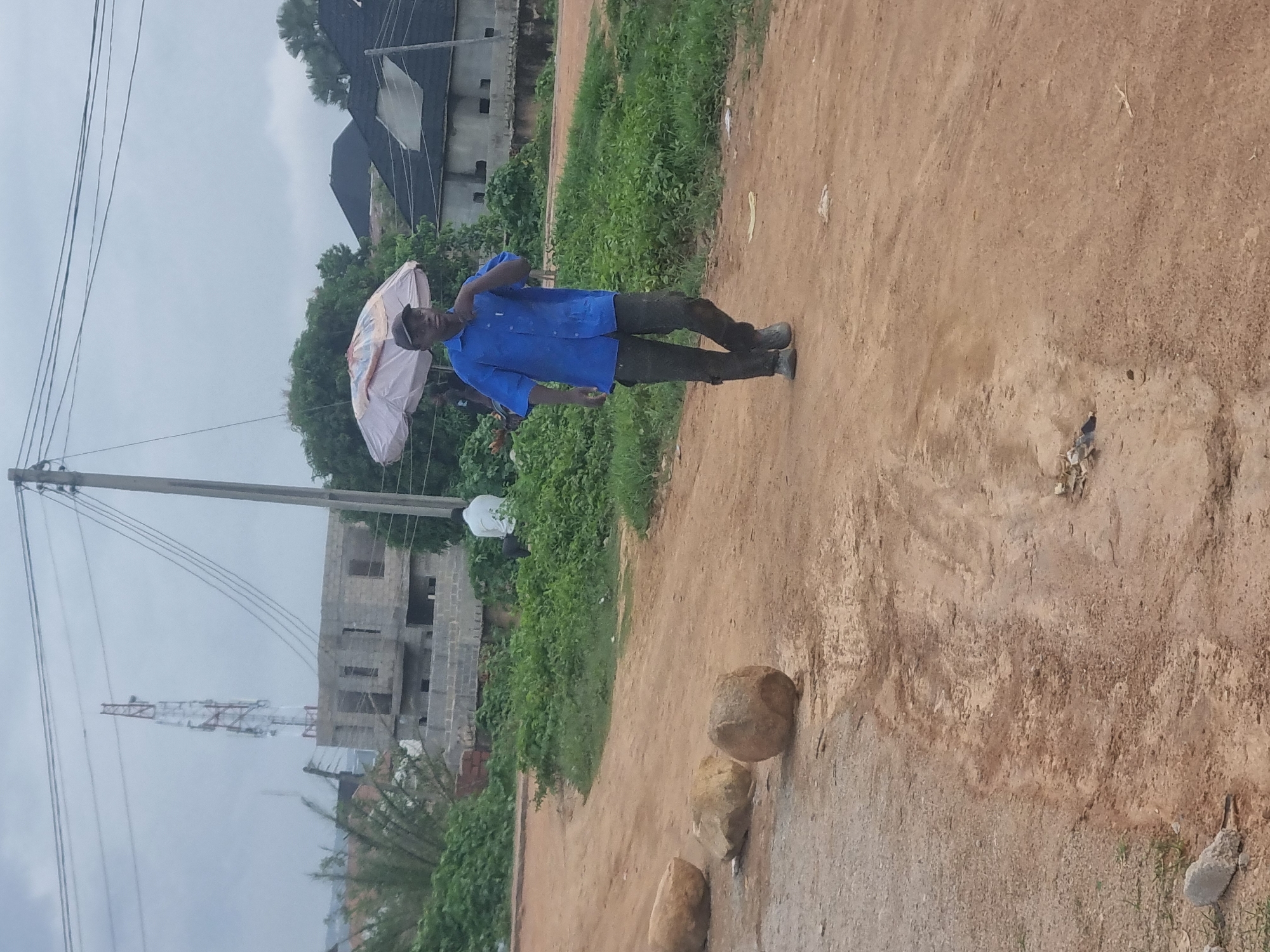 In Gwong, Nigeria, a man stands on a dusty path, holding an umbrella. He wears a blue short-sleeved shirt and dark pants, with dark shoes. The umbrella is open, suggesting it is raining or sunny. Behind him, green vegetation grows, and in the distance, unfinished buildings and a construction crane are visible. Power lines stretch across the scene, indicating a populated area. The sky is overcast, hinting at a cloudy or rainy day. The ground is a mixture of dirt and gravel. Three large stones are present on the right side of the path, with smaller stones scattered around them. The overall impression is one of a somewhat rural or developing urban environment.