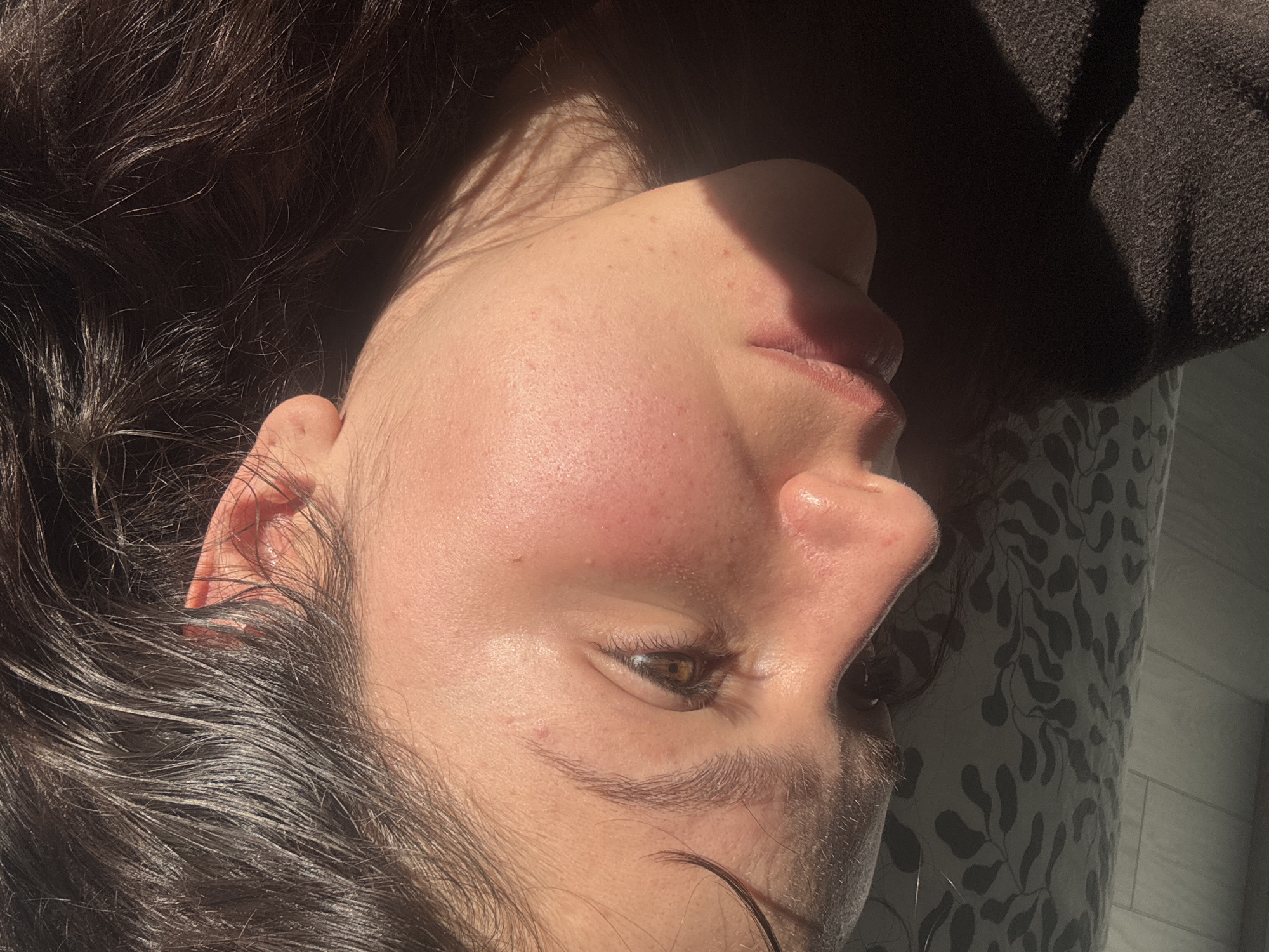 A close-up, high-angle shot captures the right side of a person's face, oriented horizontally in the frame. The subject has dark, wavy hair, and appears to be lying down, with a patterned pillow or fabric beneath their head. Strong, warm natural sunlight illuminates the face from the left, casting sharp shadows, particularly across the nose and under the chin, suggesting either early morning or late afternoon.
The person's skin tone is warm, with visible pores and a few small, reddish blemishes or freckles on the cheek and forehead area. The right eye is partially visible, featuring a brown iris and delicate eyelashes. The lips are slightly parted, revealing their natural texture. The ear is also visible, with sunlight highlighting its contours.
On the top right of the frame, a dark, textured fabric, possibly a sweater or blanket, is visible. The bottom right shows a patterned fabric, likely a pillowcase or bedsheet, with a repeating leaf or floral design in muted tones. The overall impression is one of relaxation or repose, bathed in natural light. There is no visible text in the image. The scene is set in Burgas, Bulgaria.