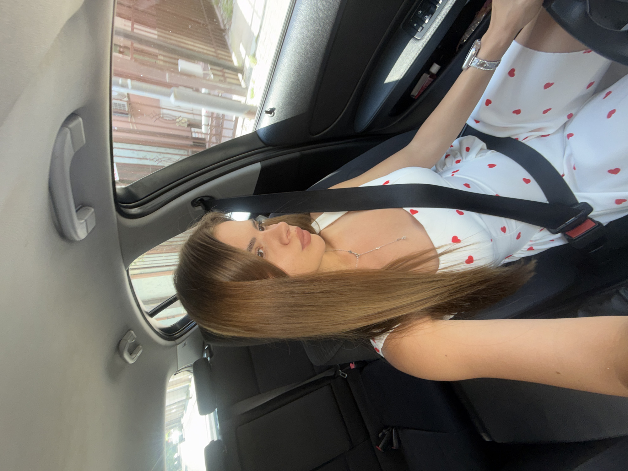 A young adult female is depicted in the driver's seat of a stationary or slow-moving vehicle in Plovdiv, Bulgaria. She is positioned facing towards her right, away from the camera, which is held to her left (passenger side). Her long, straight, brown hair, with lighter highlights, cascades over her left shoulder. She wears a white sleeveless dress or top adorned with small red heart patterns. A black seatbelt is fastened securely across her chest and lap, featuring a red and black buckle. A silver-toned necklace with a small pendant is visible, as is a silver-toned watch with a decorative, possibly diamond-encrusted, band on her left wrist.
The car's interior features a dark, likely black or dark grey, dashboard and door panels. The right-side door panel has power window controls and a storage compartment, which contains a small white bottle and a red rectangular object. A black door lock pin is visible near the top of the door. The car's headliner is light grey, with a visible grab handle above the rear passenger side. The rear seats are upholstered in black fabric and appear unoccupied.
Through the windows, an urban environment is visible. The right-side window reveals a street scene with older-style buildings, possibly with textured facades or decorative elements, and what appears to be a metal fence or gate structure. The ambient lighting suggests a bright, sunny daytime. Through the rear window, a street with indistinct structures or vehicles is visible in the distance.