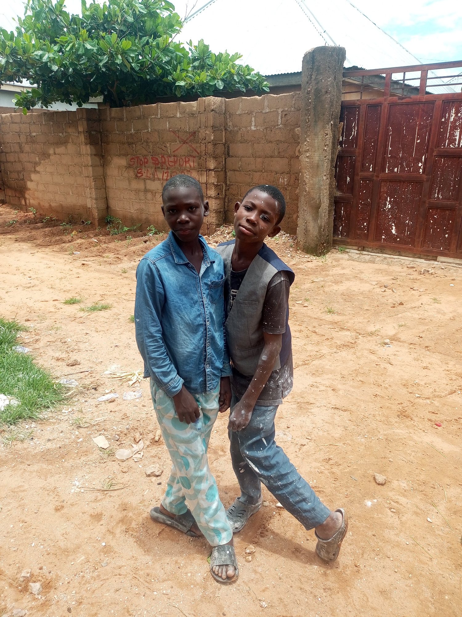 Two young boys stand outdoors on a dirt ground, with a brick wall and a metal gate in the background. The boy on the left is wearing a blue denim shirt and light-colored pants with a geometric pattern. The boy on the right is wearing a dark vest over a t-shirt and dark pants. Both boys have some white smudges on their faces and clothes, suggesting they may have been involved in some activity that produced this residue. The environment appears to be somewhat unkempt, with scattered debris on the ground and some vegetation visible. The brick wall has red graffiti on it. The metal gate is rusted and has white paint splatters. The sky above is partly cloudy. The scene is likely located in a residential area within Tempure, Nigeria.