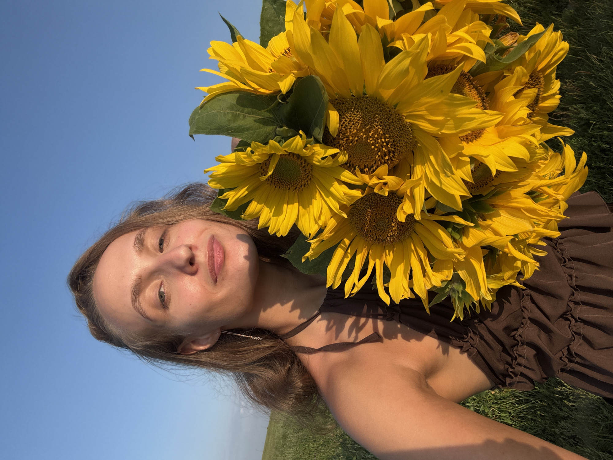 A young woman with light brown hair and fair skin stands outdoors in a field of green grass under a clear blue sky. She is holding a large bouquet of sunflowers, with their bright yellow petals and dark centers filling a significant portion of the frame. The woman is wearing a dark brown, ruffled dress. Her left shoulder and arm are visible, as is part of her chest. She is looking directly at the camera with a slight smile. The lighting suggests it is late afternoon or early evening, casting a warm glow on her skin and the sunflowers. The image appears to be a selfie taken in Aksaitovo, Russia.