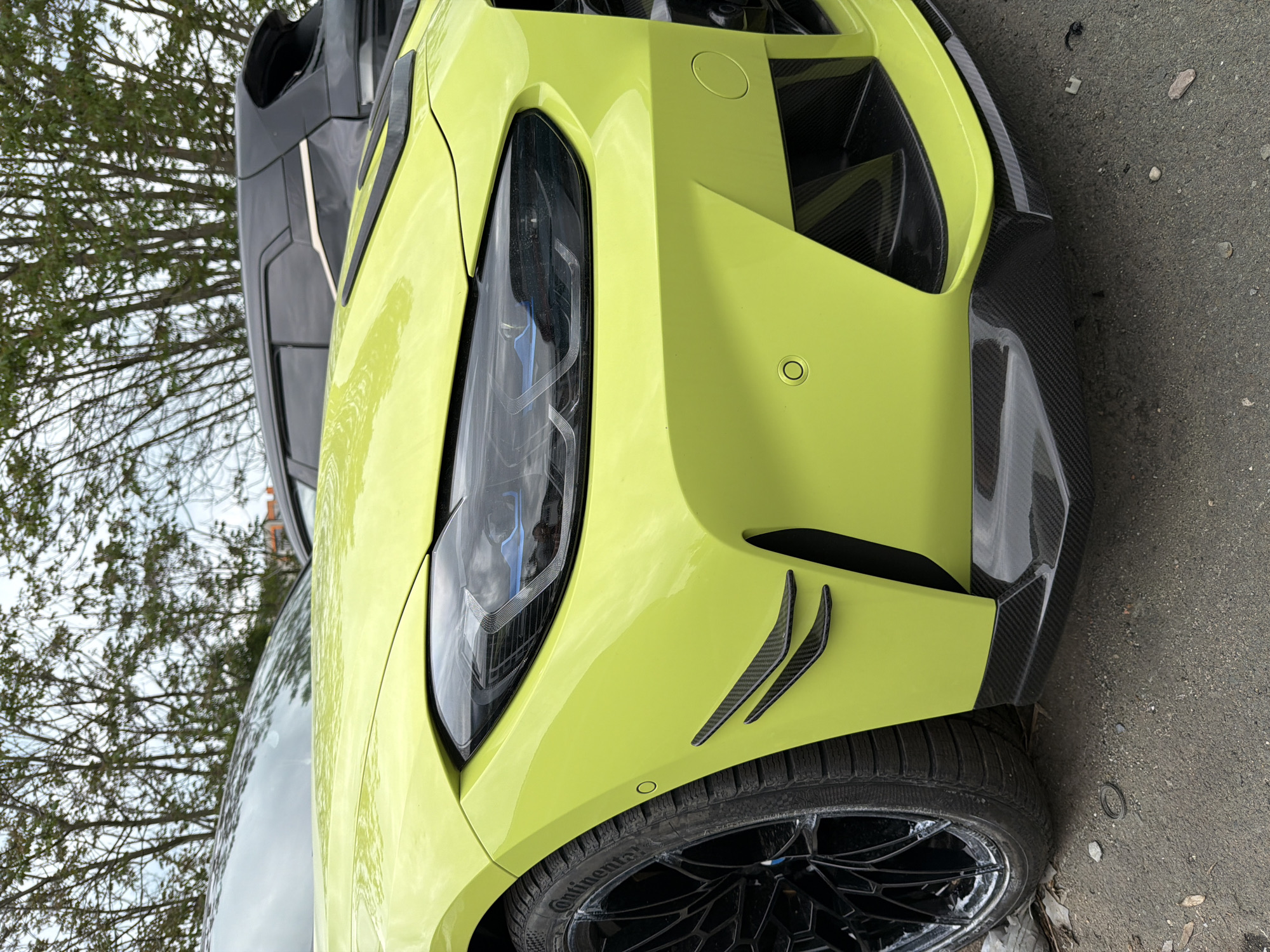 A close-up, slightly angled shot shows the front left side of a chartreuse-colored car, possibly in Burgas, Bulgaria. The vehicle features a distinctive headlight design and aerodynamic carbon fiber elements near the wheel well. A partially open, dark-colored panel is visible above the hood. The car tire is black and has a detailed multi-spoke rim design. Background elements consist of tree branches and a glimpse of the urban environment. The car is parked on a gray concrete surface.