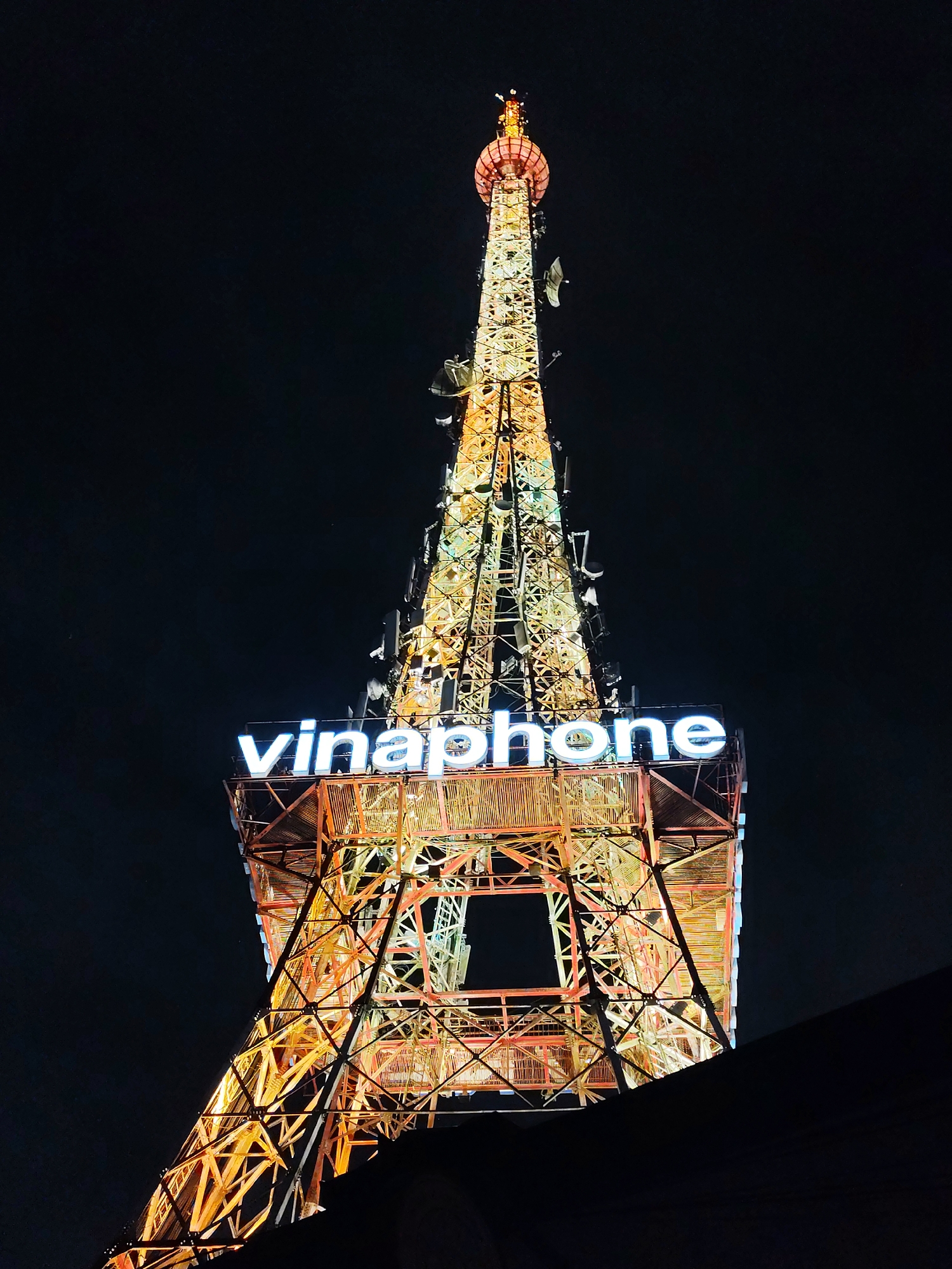 The image depicts the Vinaphone TV Tower at night in Da Lat, Vietnam. The tower is illuminated with a golden-orange light, highlighting its metal structure and supporting numerous antennas. The "vinaphone" signage is prominently displayed in bright white neon lights. The background is a dark night sky. A portion of a dark, shadowed structure is visible at the bottom of the frame.