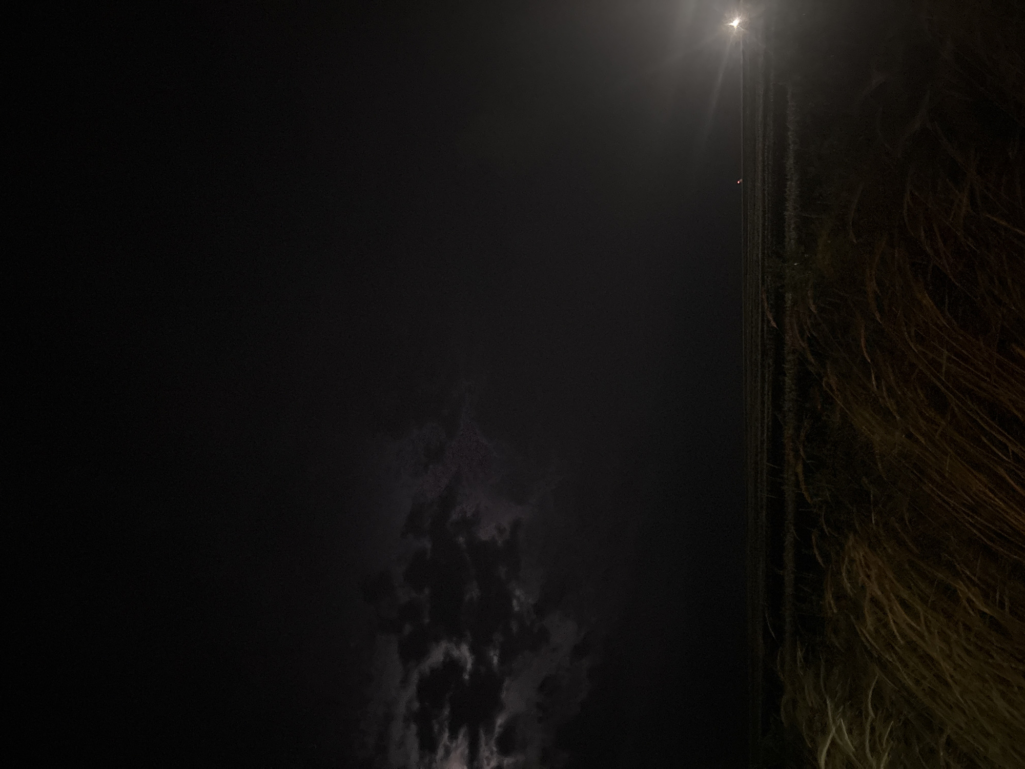 The image depicts a nighttime scene in Burgas, Bulgaria. The frame is divided unevenly, with the left side being predominantly dark, suggesting a low-lit area or a body of water, potentially the Black Sea. A diffuse glow, likely from the moon, is visible at the top, illuminating a section of the sky. Irregular cloud formations are discernible in the lower central area, exhibiting varying shades of gray and providing a sense of depth. The right side of the image contains a dense mass of what appears to be long hair, its color a blend of dark brown and lighter highlights, obscuring details of the background. A faint, vertical line suggests a street or a path alongside the hair, perhaps illuminated by distant light sources.
