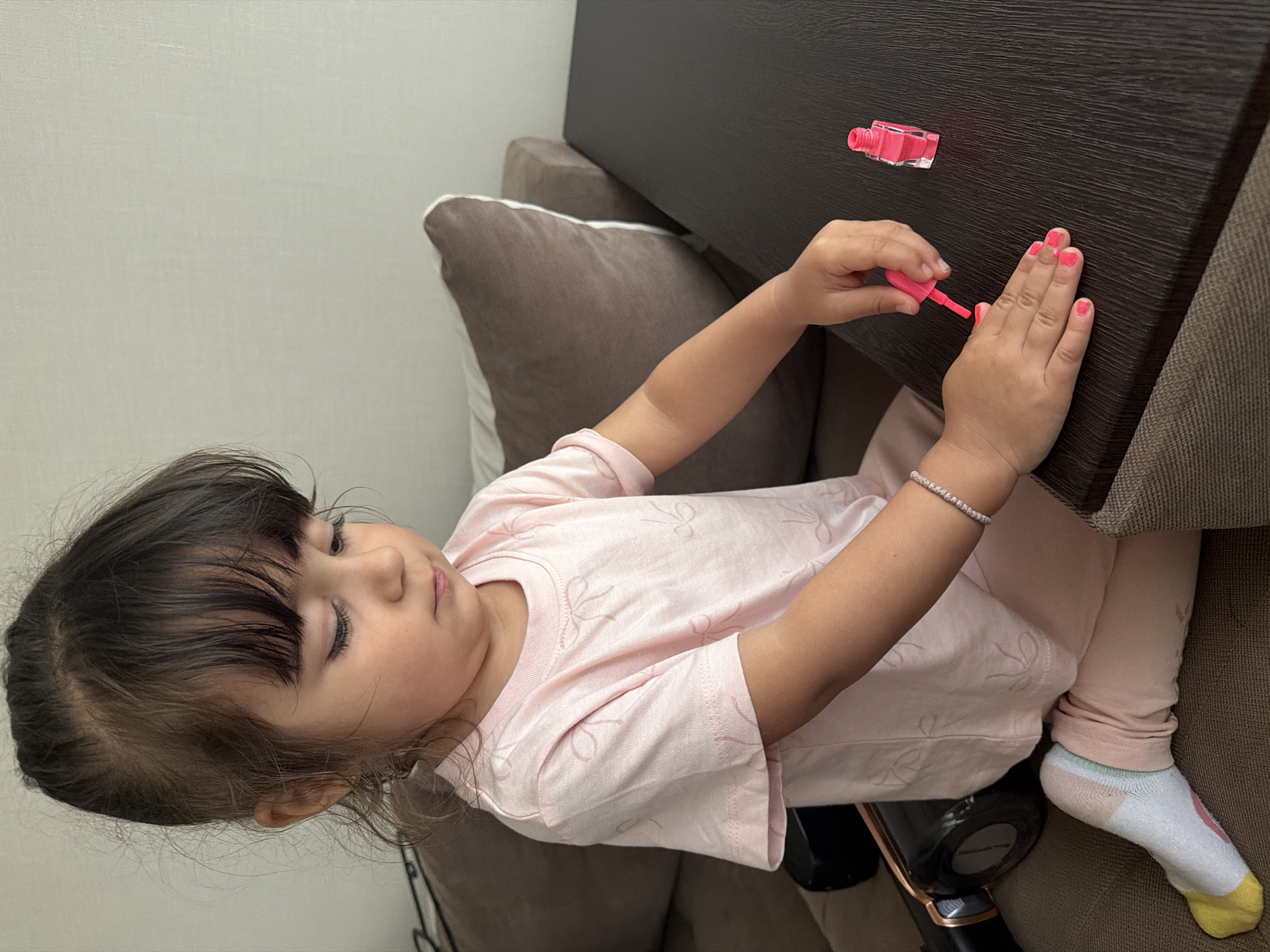 A young girl is depicted indoors, likely in a living room in Burgas, Bulgaria, sitting on a brown sofa next to a dark brown table. She is actively engaged in painting her fingernails with bright pink nail polish.
The girl, appearing to be a toddler or preschooler, has dark hair with bangs and is dressed in a light pink t-shirt adorned with subtle bow patterns, paired with light pink leggings. Her left foot, wearing a white sock with a yellow toe and green/pink stripes, is visible. A silver-colored beaded bracelet adorns her left wrist.
She holds an open bottle of pink nail polish with a brush in her left hand, concentrating intently on applying the polish to the nails of her right hand, some of which are already painted pink. Another closed bottle of the same pink nail polish rests on the table beside her. Her expression is focused and calm as she performs the activity. The background features a plain, light-colored wall. A black and bronze household appliance, with the brand "Rowenta" partially visible, is seen near her feet at the bottom of the frame. The scene captures a moment of playful self-grooming.