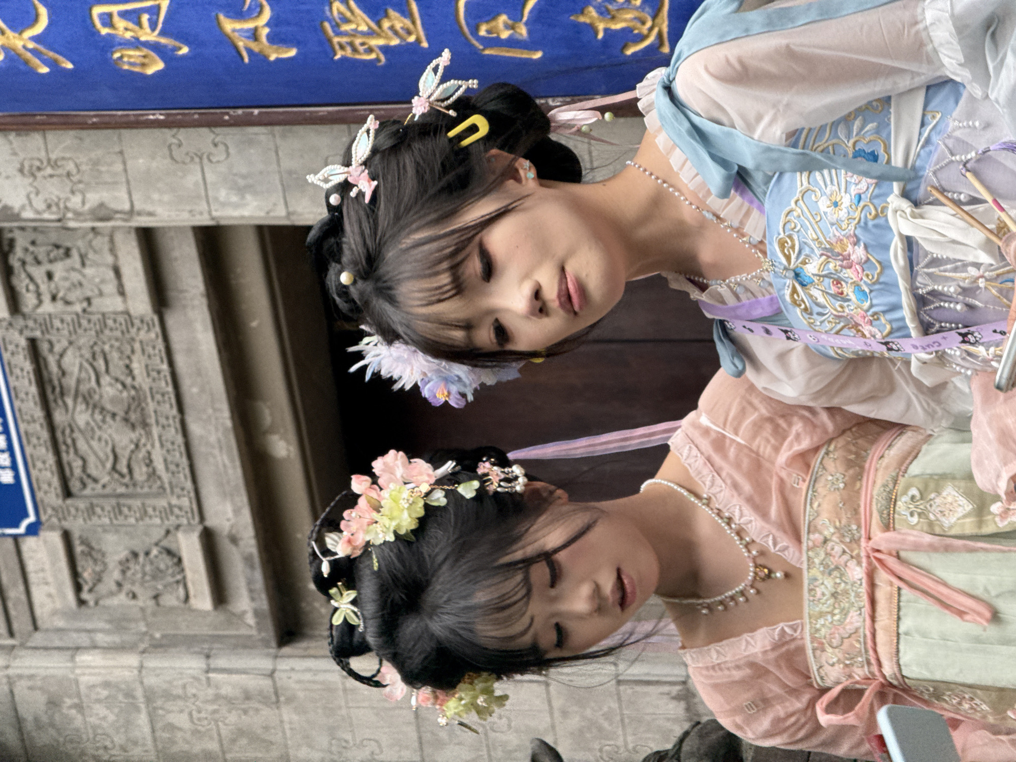 Two young women wearing traditional hanfu clothing are posing for a photo in front of an ornate stone building. They are both dressed in elaborate hairstyles with flower and butterfly ornaments, and pearl necklaces. One woman is wearing a blue and white hanfu, while the other is wearing a pink and green hanfu. The building has intricate carvings and a blue sign with gold lettering. The location is likely in Yuzhong District, Chongqing, China.