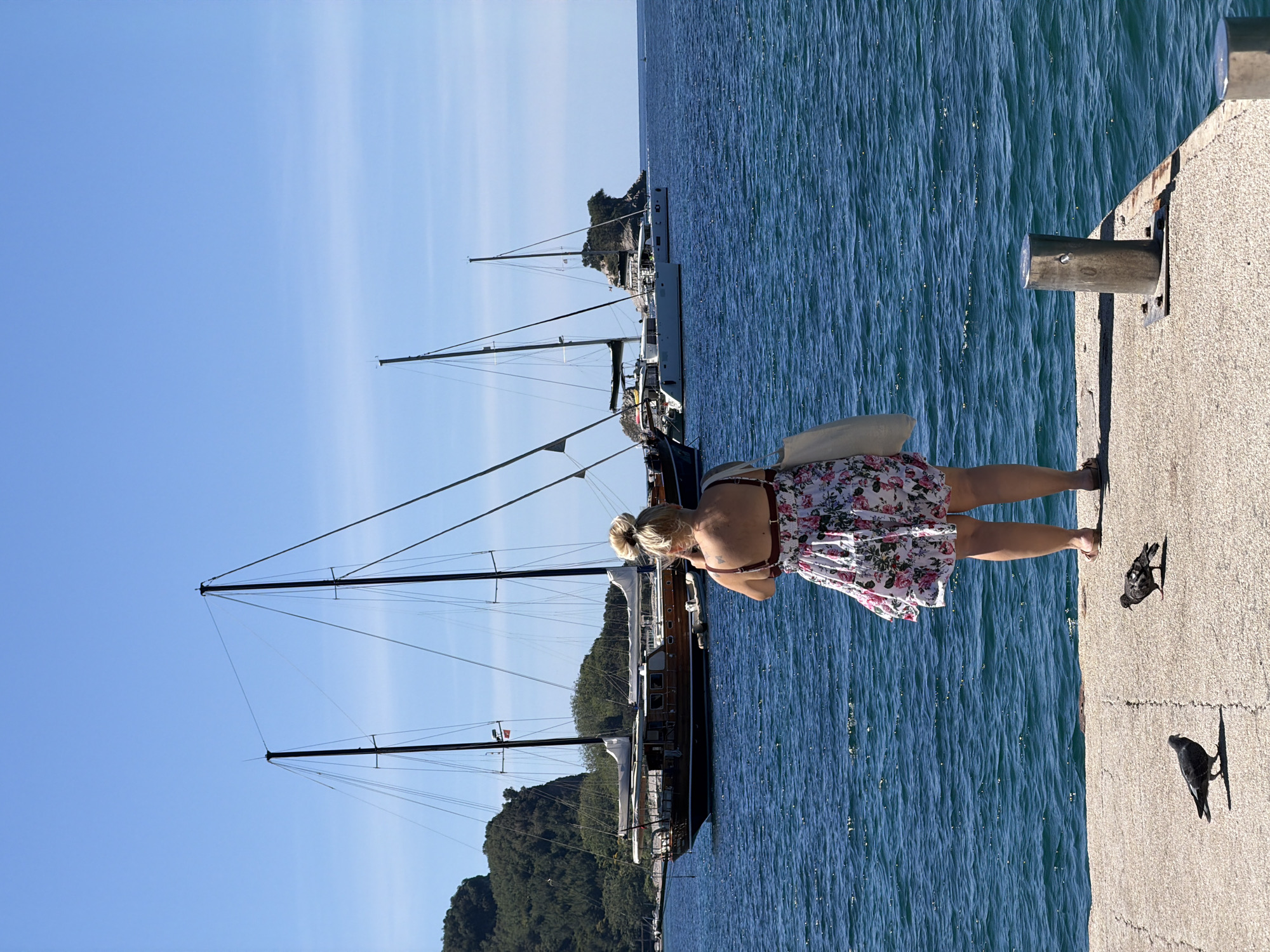 A woman in a floral dress stands on a concrete pier near the Adriatic Sea in Budva, Montenegro. She is looking down toward the water, near where two dark birds stand on the pier surface. Moored sailboats with tall masts can be seen in the background, along with green, hilly terrain. The sky is bright blue.