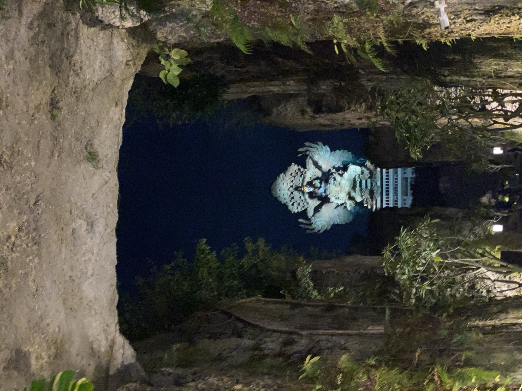 The image depicts a nighttime view of the Garuda Wisnu Kencana (GWK) Cultural Park in Ungasan, Kuta Selatan, Bali, Indonesia. A colossal statue, believed to be of Vishnu riding Garuda, is illuminated against the dark sky, seen through an archway formed by rocky cliffs. The statue is predominantly blue and white, with large, spread wings. Vegetation, including trees and ferns, grows on the rocky walls of the archway. The ground below is dimly lit by scattered lights, suggesting a pathway or viewing area. The scene is dramatic and awe-inspiring, highlighting the scale of the statue and the natural rock formations.