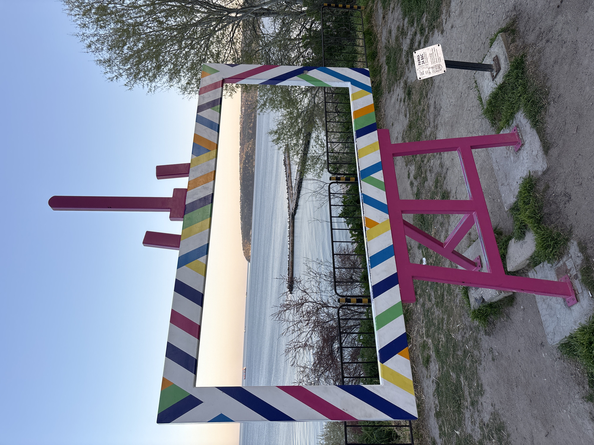 The image depicts a large, rectangular frame constructed to resemble a director's clapboard, positioned outdoors in Varna, Bulgaria. The frame is painted with colorful diagonal stripes of white, blue, green, yellow, orange, and pink. It is supported by a pink, easel-like structure. The frame captures a view of the sea, a breakwater, and the coastline. A small sign is attached to a post next to the structure. The ground is gravel and dirt, with some patches of grass. Trees are visible in the background and within the framed view.