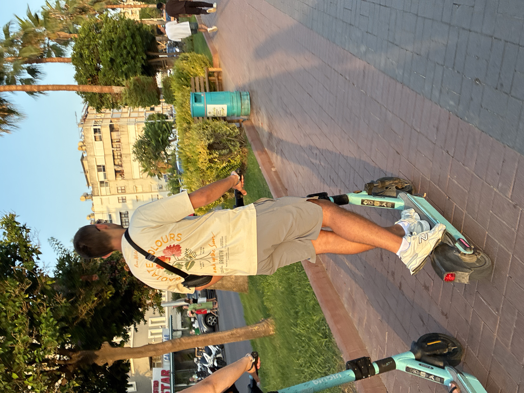 In Alanya, Turkey, a man rides a light blue electric scooter along a brick-paved walkway lined with green bushes and palm trees. He is wearing a light-colored t-shirt with a floral design and text that includes "GROWTH," gray shorts, and white sneakers. A black crossbody bag is slung over his shoulder. Another scooter is visible to his left. In the background, buildings and parked motorcycles are present. The scene is illuminated by the warm light of a setting sun.