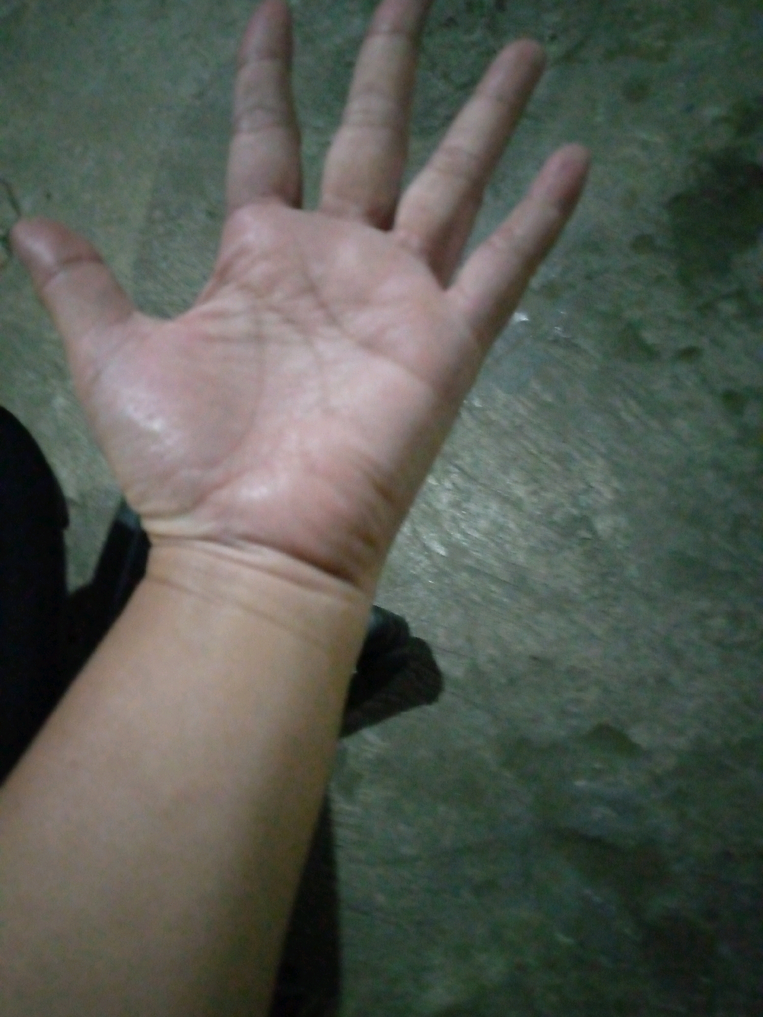 A close-up, low-angle shot shows a left hand and forearm against a dark, textured surface. The hand is open, palm facing upwards, with fingers extended. The skin appears light-skinned and slightly moist. The forearm is also visible, extending downwards and appearing smooth. The background is a dark, grayish-green, possibly concrete or asphalt, with some lighter patches and texture. There is a dark shadow visible in the lower left portion of the image. No specific landmarks or elements that would place the image in Sumberwono, Indonesia, are present.