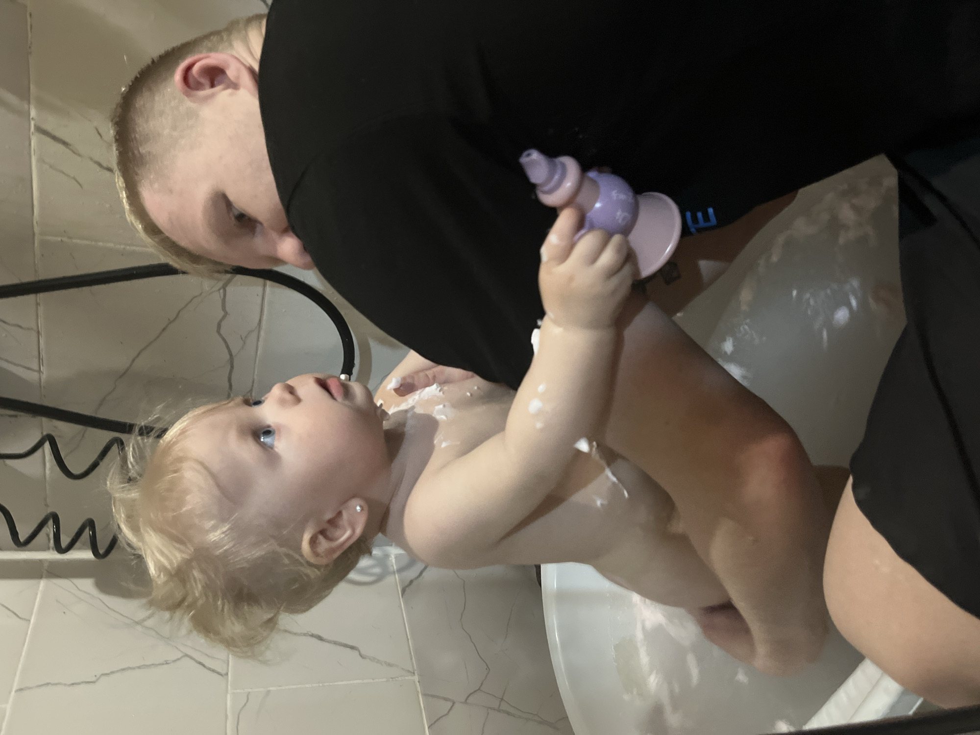 A fair-skinned adult male in a black t-shirt is holding a fair-skinned baby in a white bathtub, likely in a bathroom setting. The baby is partially covered in white foam, possibly soap. The baby is holding a small purple and pink toy. The bathroom features tiled walls with a grey marbled pattern. A black metal rack is attached to the wall. The location of the scene is the city of Vesyoloye, Russia.