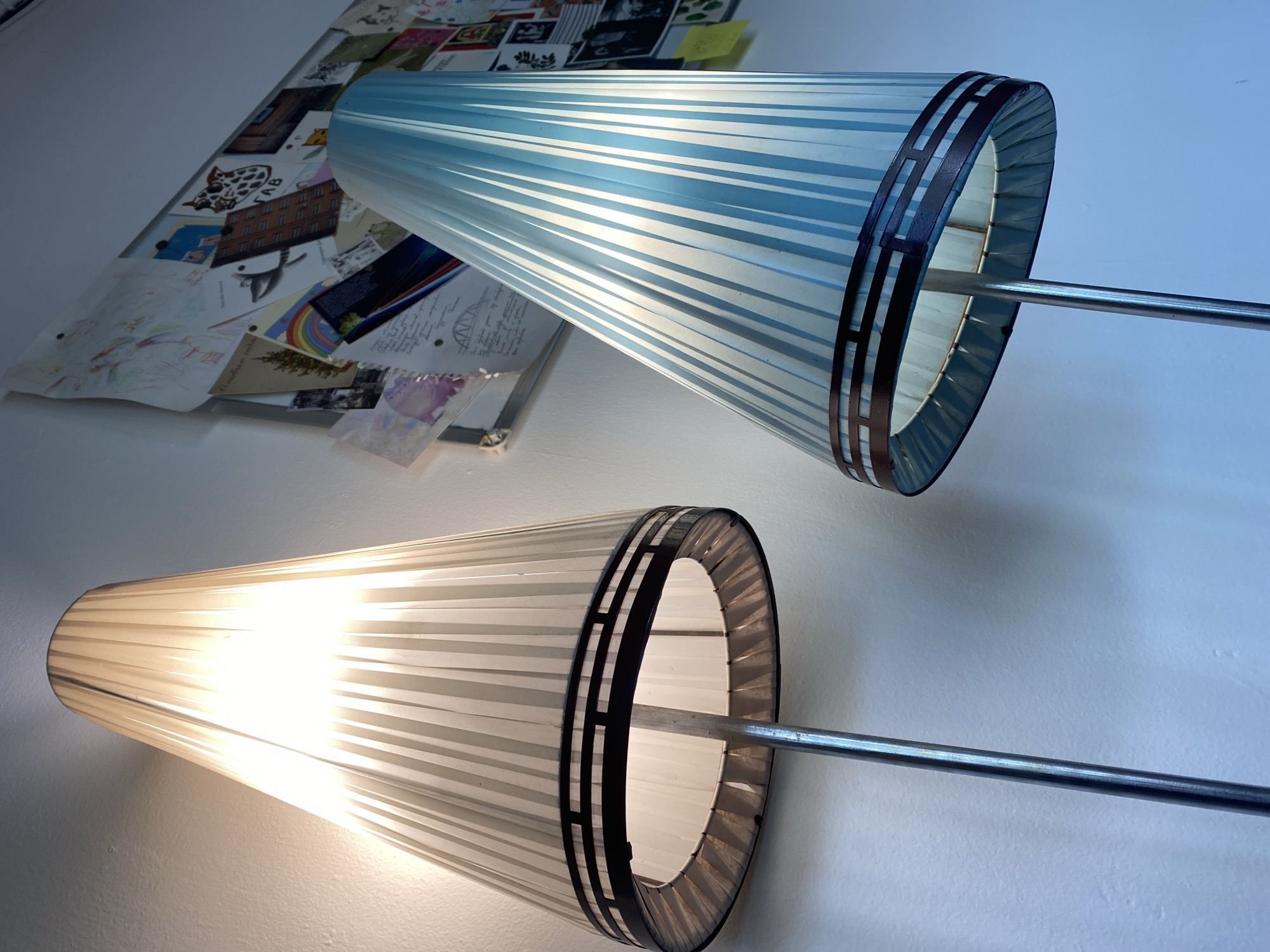 Two illuminated lamps with striped shades are affixed to a wall. The top lamp has a blue and white striped shade, while the bottom lamp's shade is white. A collage of images and papers is mounted on the wall above the blue lamp. The lamps are mounted on metal rods. The lighting suggests an indoor setting. The image does not depict Rusanovo or provide information about it.