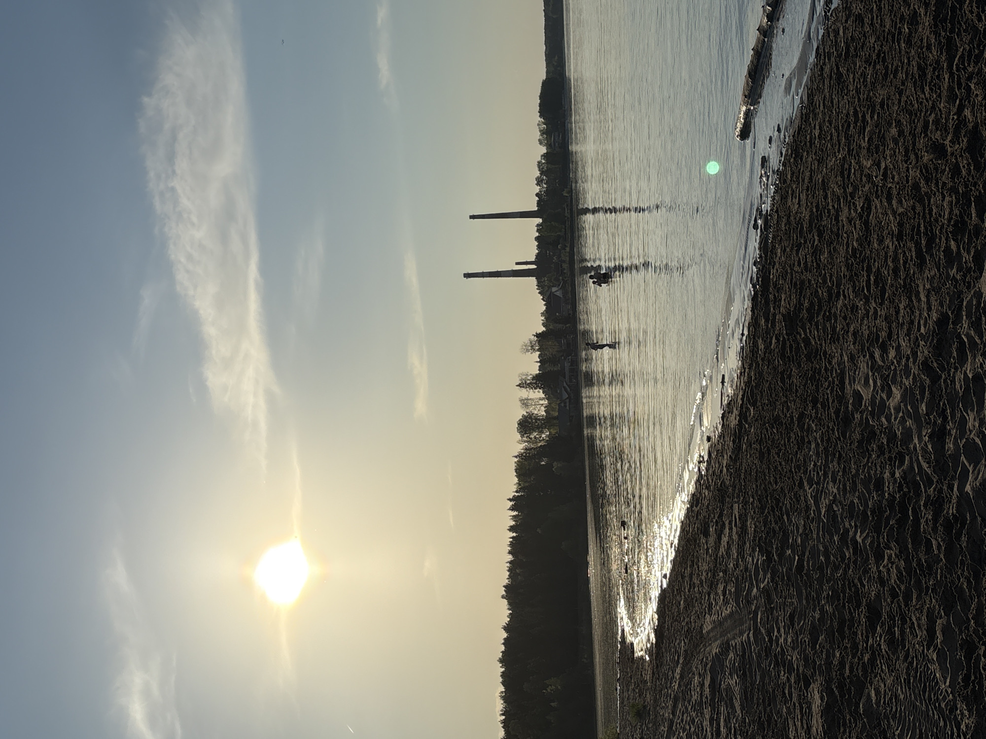 The image shows a landscape at dusk in Chaikovsky, Russia. The scene includes a body of water, likely a lake or river, with a sandy shore in the foreground. Two tall, slender industrial chimneys are visible in the distance on the opposite side of the water, indicating an industrial area. The sun, low on the horizon, is emitting a bright glow in the sky, creating a glare. The sky is mostly clear with some scattered clouds. Trees are visible along the shoreline, and there may be a boat or small vessel on the water. The sand on the shore appears to be textured.