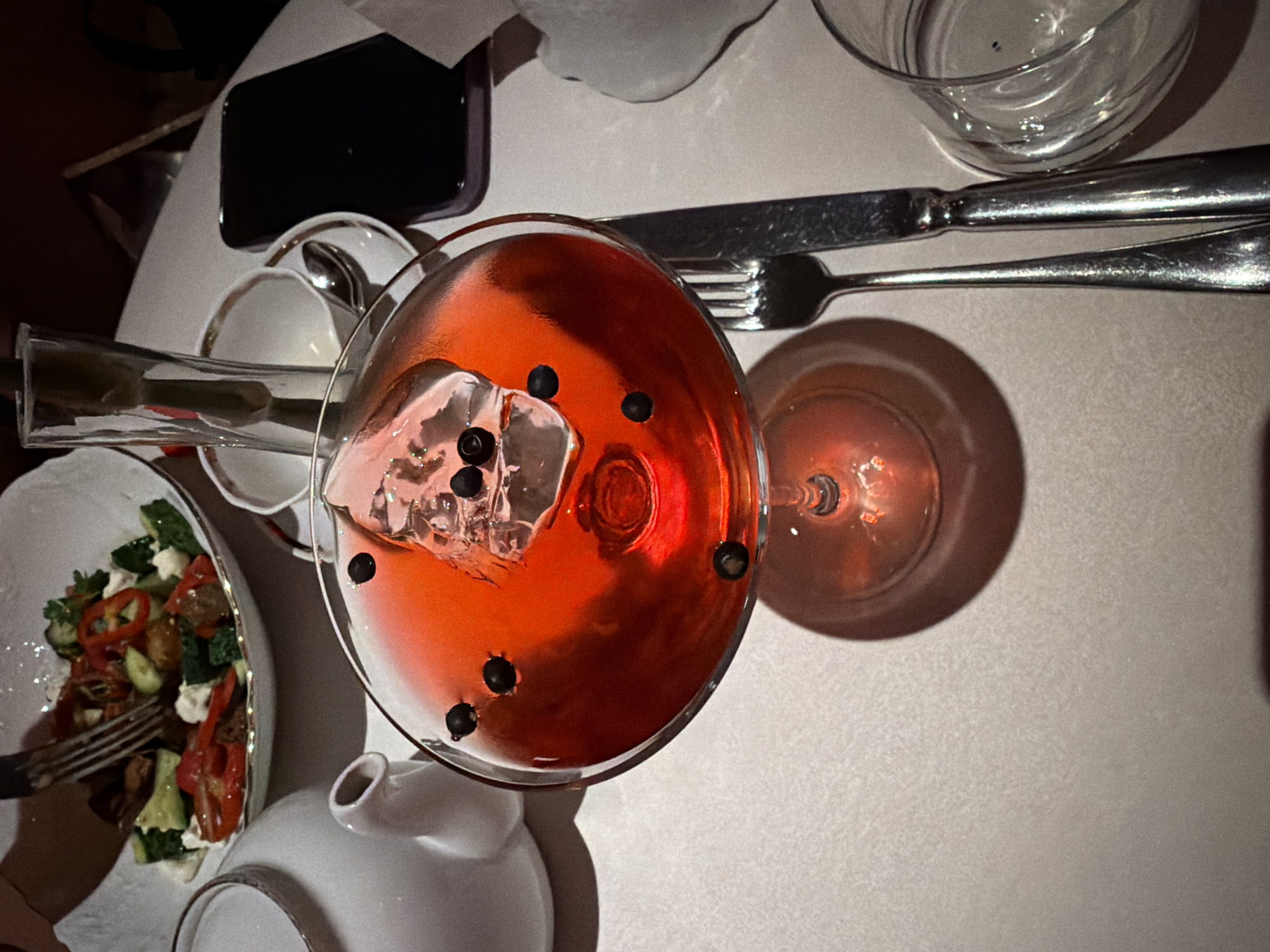 The image shows a high-angle view of a table setting, likely in a restaurant. On the table, there's a salad in a white bowl with a fork partially visible within, a red drink in a martini glass with ice and black berries, a small white teapot, a clear glass, a knife and fork, and a black cell phone. There is also a small glass vase with flowers. The color palette is dominated by reds, whites, and silvers. The lighting appears soft, creating gentle shadows. The scene depicts a meal setting with drinks and salad. There is no verifiable information in the image to confirm that the location is Moscow, Russia.