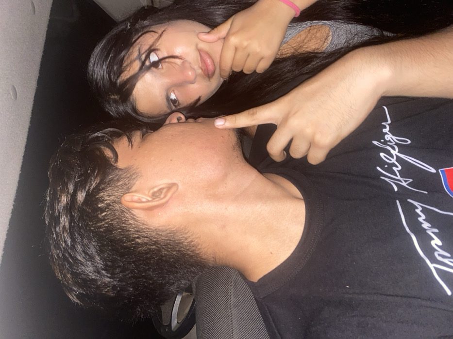 A young couple poses closely. The man, wearing a black Tommy Hilfiger shirt, reclines with his head tilted back. A woman with long, dark hair lies above him, her face turned to the camera. Her finger touches his cheek. She wears a gray top and a pink wristband. The setting appears to be indoors, with dark tones and a hint of what may be a car seat visible in the lower left corner.
