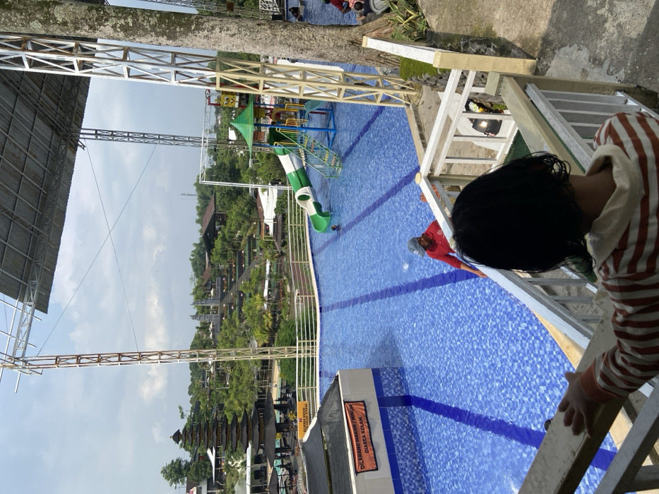 A child looks down from a stairway toward a blue-tiled swimming pool. In the pool, a person in red stands near the edge. A water slide is positioned on the opposite side of the pool near a playground structure. Trees and buildings line the background. A sign is posted on the near side of the pool. Metallic structures appear overhead.