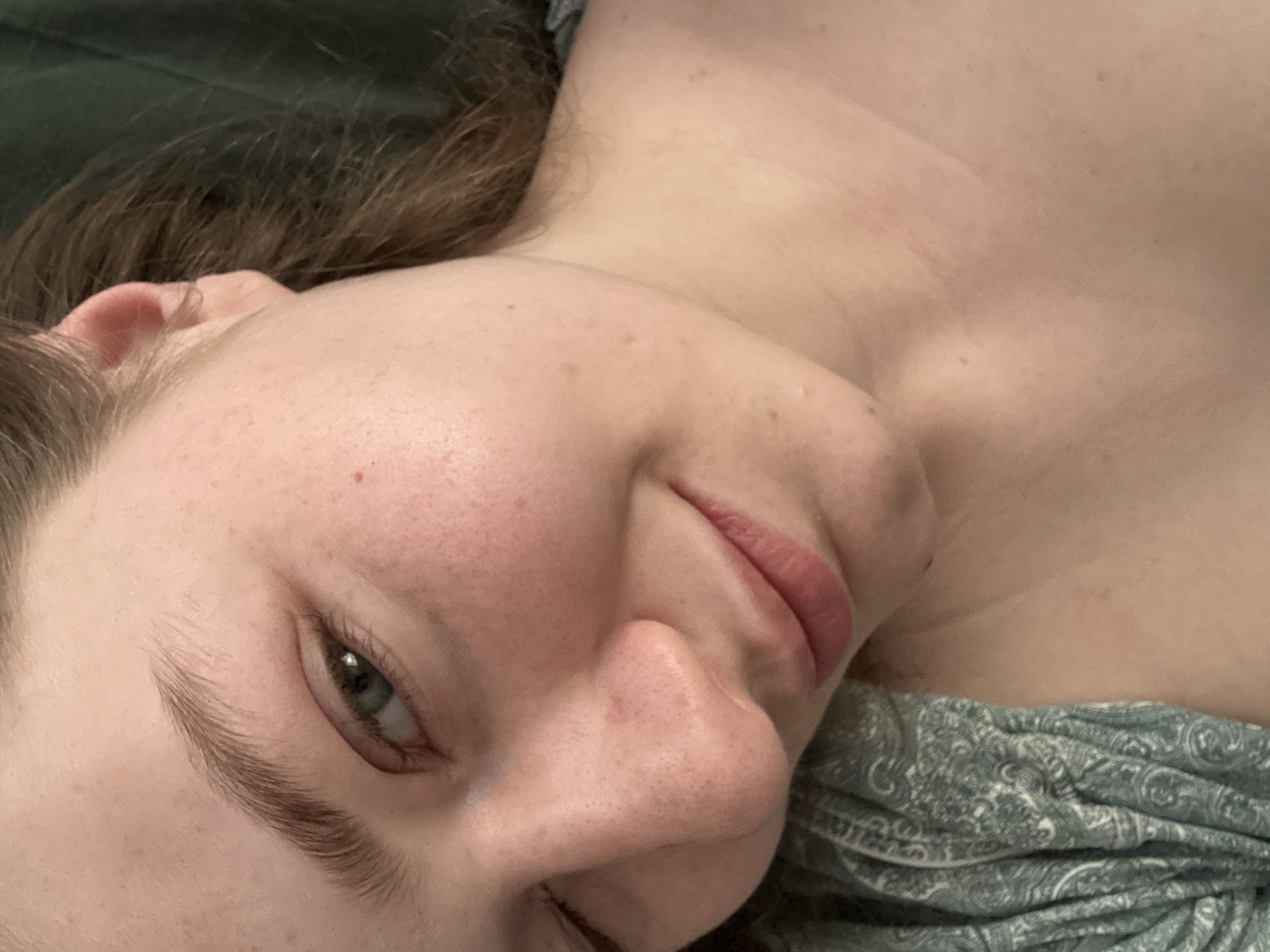 A close-up, angled portrait shows a light-skinned person with green eyes and light brown hair. They are lying down, with their face tilted towards the camera. A paisley patterned fabric is visible under the person's neck. The background is blurred. There is no information in the image to indicate the location is Kazan, Russia.