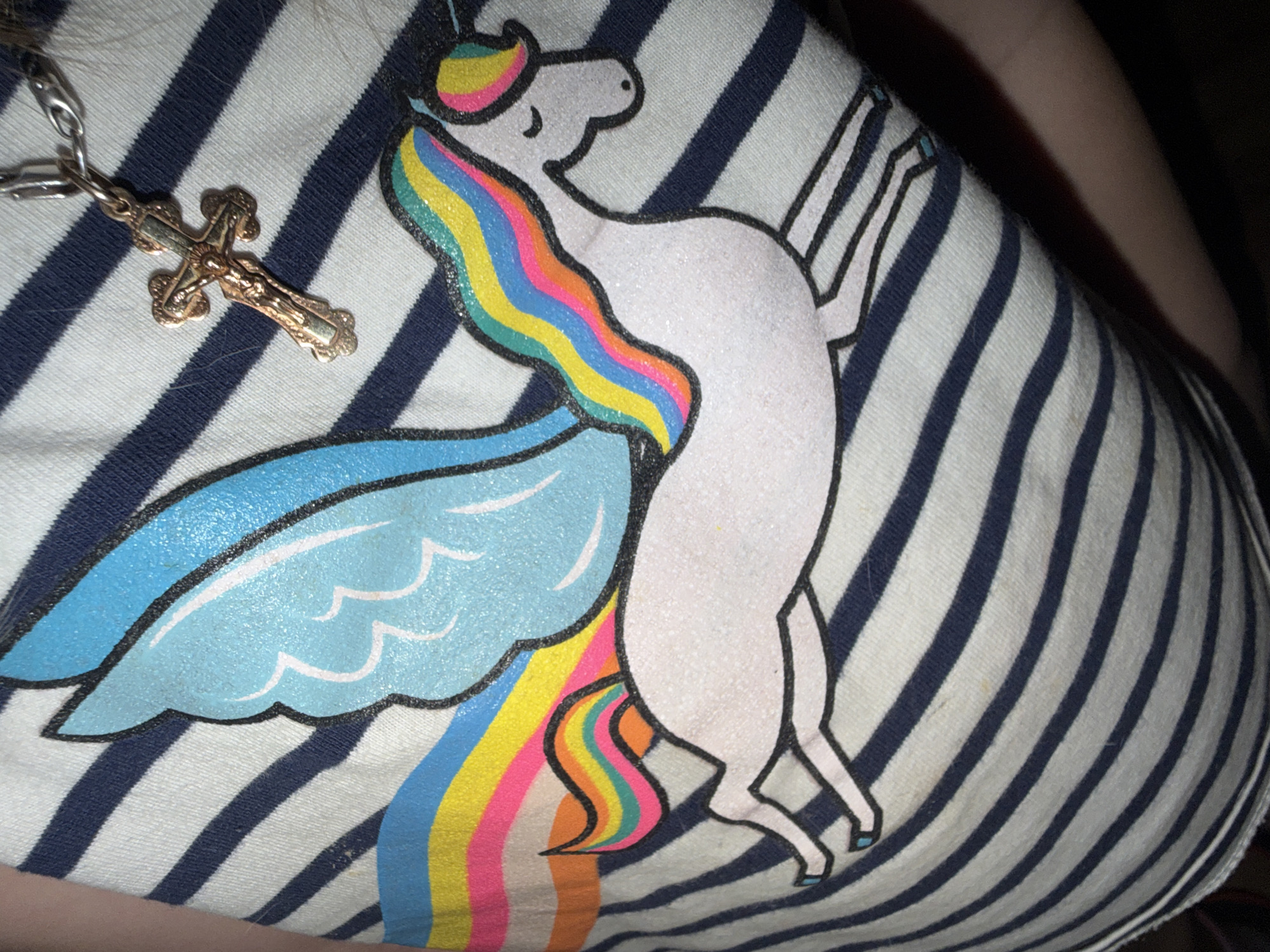A close-up, overhead, medium shot shows a person wearing a horizontally striped shirt with a graphic of a winged unicorn with a rainbow mane and tail. A gold-colored crucifix pendant on a silver chain rests on top of the shirt, near the unicorn's head. The shirt is white with navy blue stripes. The unicorn graphic is white with a light blue wing, and the rainbow colors are pink, yellow, orange, green, and blue. The image is taken from an unknown location in Unknown, Unknown.