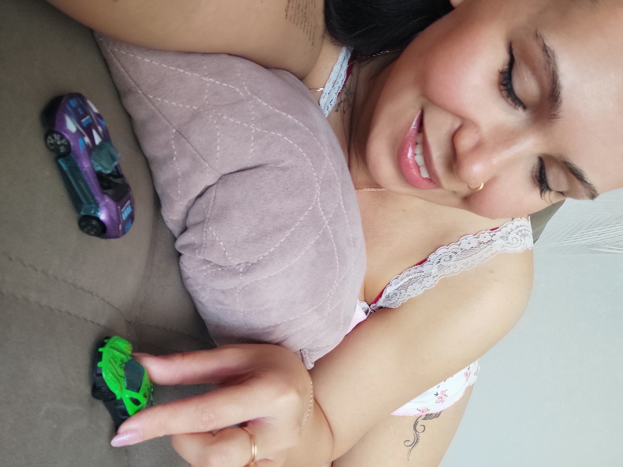 A woman is seen from an overhead perspective, resting on a brown sofa, with a mauve quilted pillow under her head. She is smiling with her eyes closed. She is wearing a lacy white and floral bra. A purple toy car lies to the left of her head. Her right hand holds a green toy car near the bottom of the image. Her hand has a gold ring on the third finger and a gold bracelet on her wrist. A tattoo is visible on her chest near her left shoulder, and another is visible on her left side. The location is Chaikovsky, Russia.