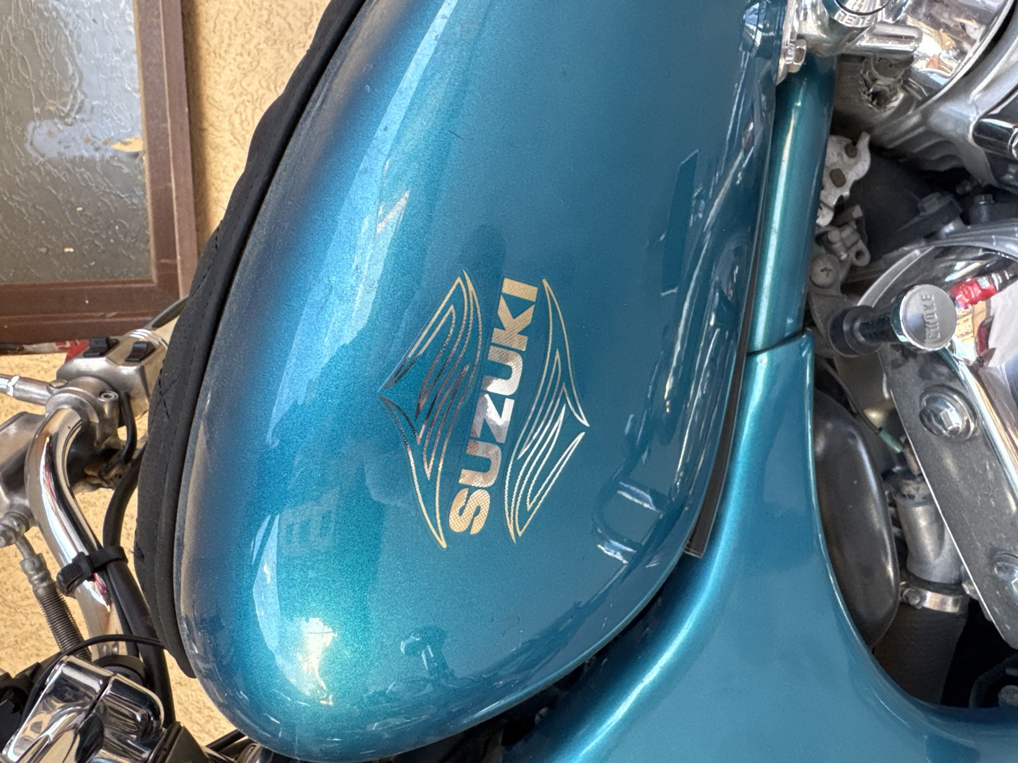 The image shows a close-up of a teal Suzuki motorcycle. The "Suzuki" logo is prominently displayed in gold with stylized wings on the fuel tank. Parts of the handlebar and engine components are visible. The photograph appears to be taken outdoors. There is no indication of Plovdiv, Bulgaria.