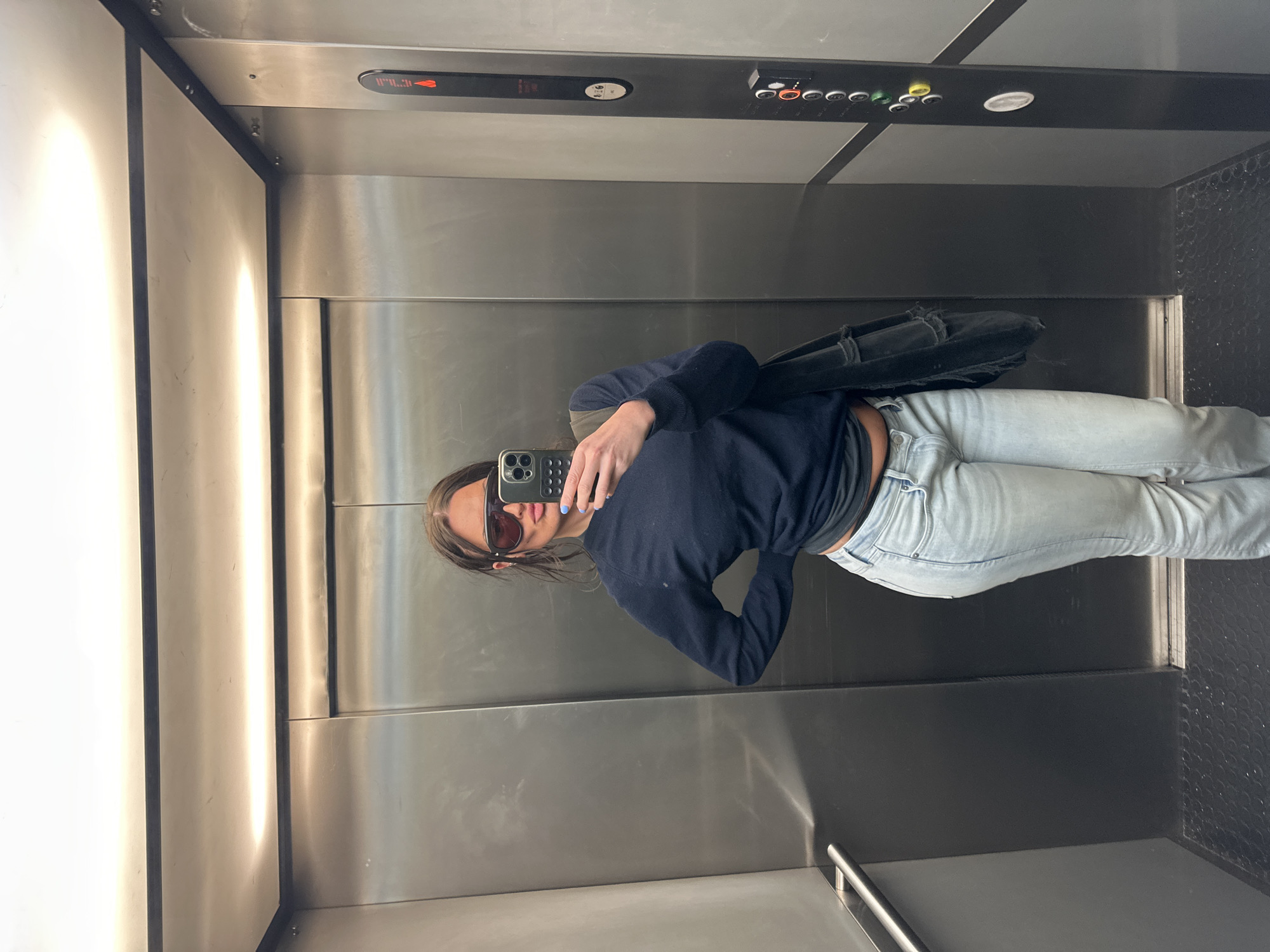 The image shows a person taking a selfie in an elevator. The elevator has stainless steel walls and ceiling. There is a panel with buttons and a digital display above the person's head. The person is wearing sunglasses, a dark blue sweater, and light-wash jeans. They are holding a smartphone in their left hand to take the picture. A black bag is slung over their shoulder. The floor of the elevator is dark gray with a textured pattern in the corner. Based on the elevator design and the presence of digital displays, the location is potentially a modern building in a city. The exact location within London cannot be determined without further context.