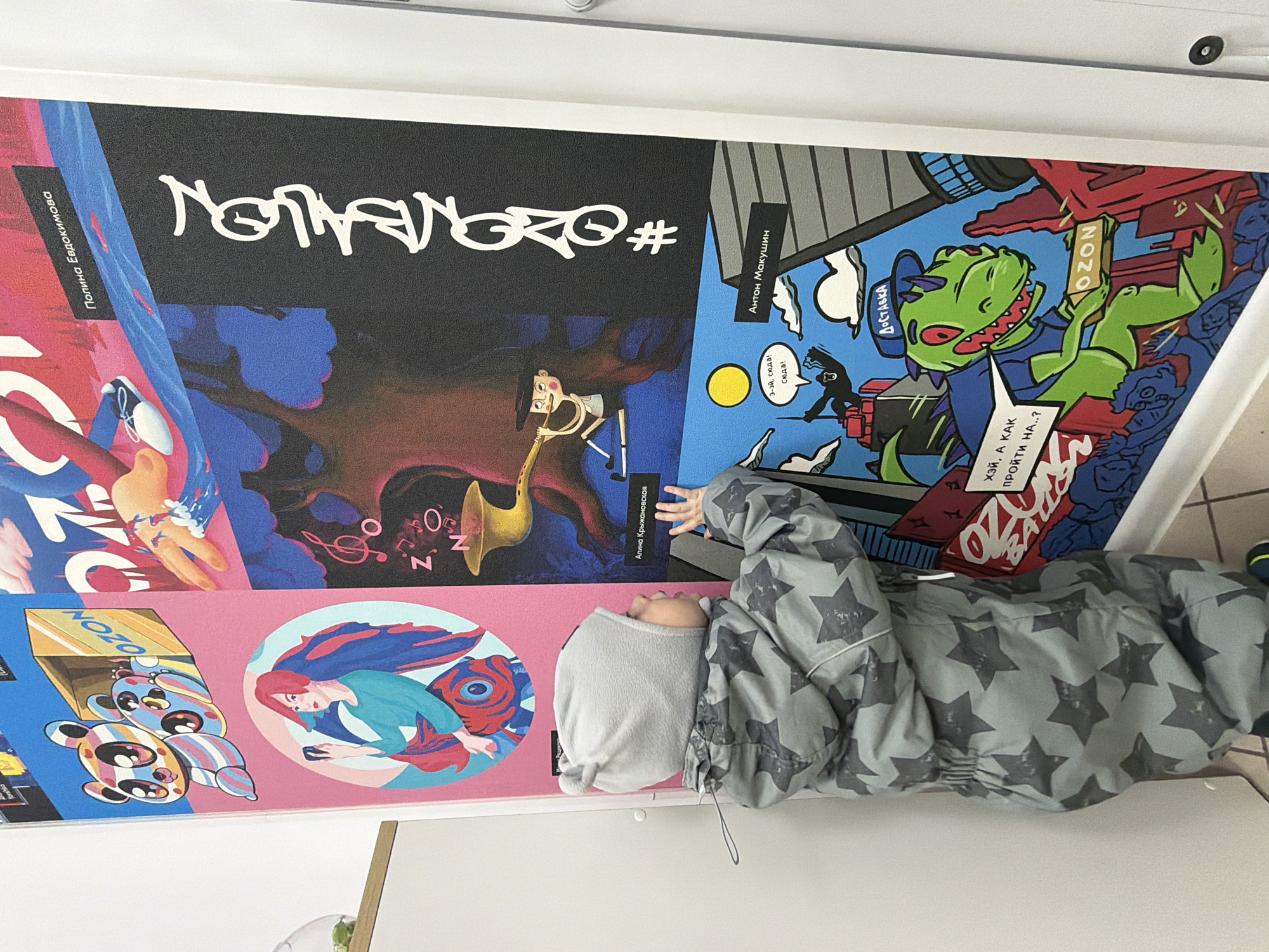 A small child, dressed in a grey star-patterned jacket or snowsuit and a light grey hood, is reaching upwards towards a large, colorful poster or mural on a white wall. The child's face is mostly obscured, but they appear engaged with the artwork. Below the poster is a flat, white surface, possibly a table or floor.
The prominent poster features a vibrant collection of cartoon-style illustrations, possibly depicting different characters or scenes related to the Russian e-commerce company Ozon. A large, stylized white text on a black background reads "#OZONBALLON".
Key visual elements on the poster include:
- A panel with a character playing a saxophone amidst musical notes.
- Another panel features a green, dinosaur-like character wearing a blue hat with "ДОСТАВКА" (Delivery) written on it, holding a brown box labeled "OZON". This character has a speech bubble saying "ХЭЙ, А КАК ПРОЙТИ НА..?" (Hey, how do I get to..?).
- A different section shows a character resembling a person with red hair, holding a phone, set within a circular frame with swirling blue and red patterns.
- Another illustration depicts colorful, segmented bear-like characters emerging from an "OZON" box.
- Additional names like "Полина Евдокимова," "Антон Макушин," and "Алина Крыжановская" are visible, likely crediting the artists.
- A speech bubble from another character says "3-эй, сюда! Сюда!" (Hey, over here! Over here!).
The scene is an indoor setting in Votkinsk, Russia, with the child interacting with this artistic display. The time of day and weather are not discernible.