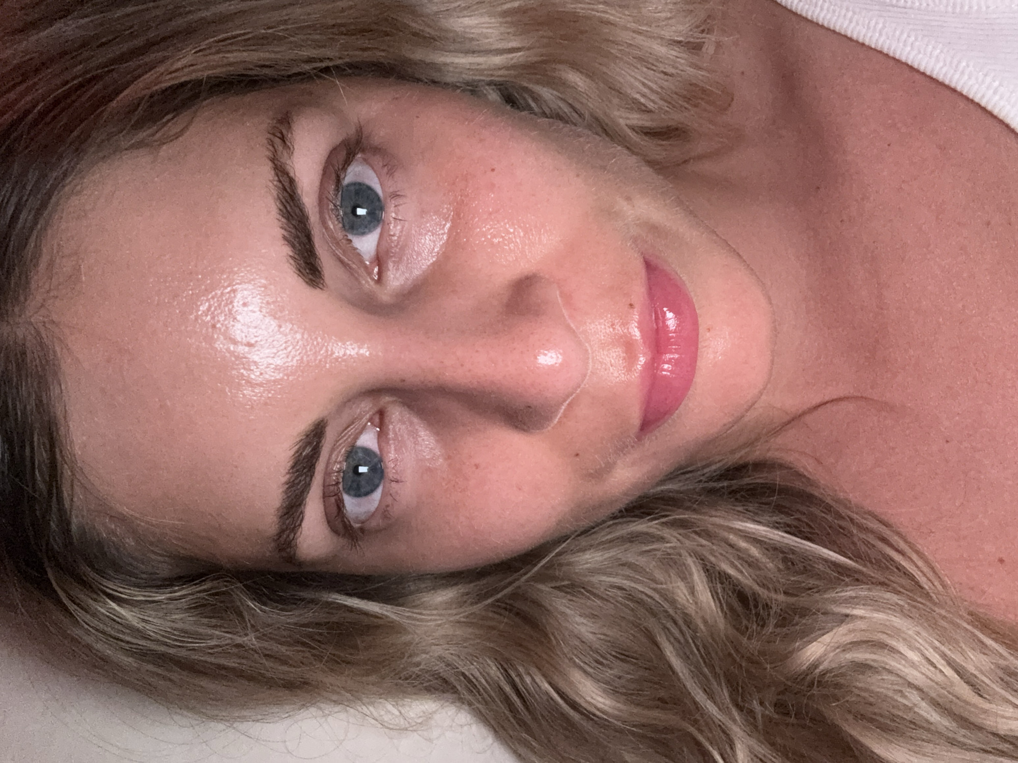 A close-up, head-and-shoulders shot of a woman with fair skin, light brown wavy hair, and blue eyes. She is looking directly at the camera. Her skin appears slightly dewy and there is a hint of pink blush on her cheeks. She is wearing pink lipstick. Her hair is parted on her left and frames the left side of her face. She is wearing a white top that is visible at the bottom of the frame. The background is a plain, light-colored wall. The image was taken indoors, likely in a residential setting, and no specific location in Paris, France, can be identified from the visual information provided.