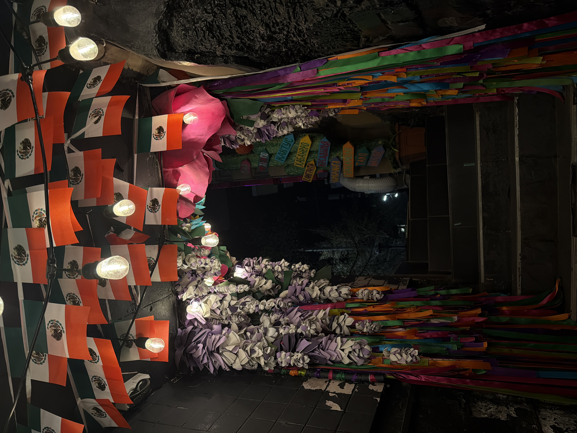 The image shows an entranceway or passageway decorated with festive elements, likely in celebration of Mexican culture. There are strings of Mexican flags with lights attached to them, along with colorful paper streamers and paper flower garlands, creating a vibrant and decorative scene. There are also small signs with the names of Mexican dishes. The exact location and date are not identifiable in the image.