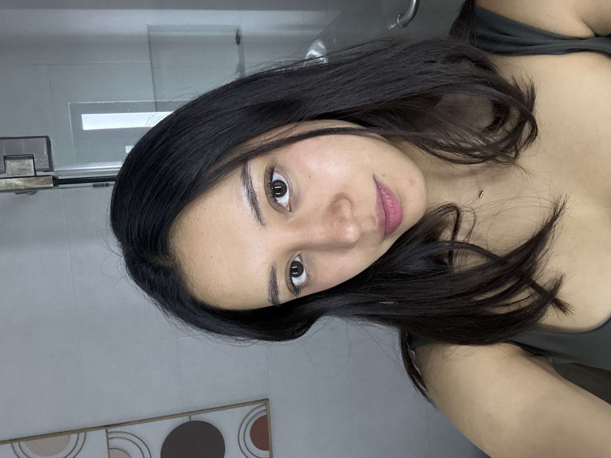 A young woman with dark brown hair is shown in a selfie taken in a bathroom. Her hair is parted to the side and frames her face. She has brown eyes, fair skin, and pink lips. She is wearing a dark-colored top. The background includes a white tiled wall, a glass shower door, and a decorative wall panel with circular designs in brown, tan, and white. The location is identified as Puebla, Mexico.