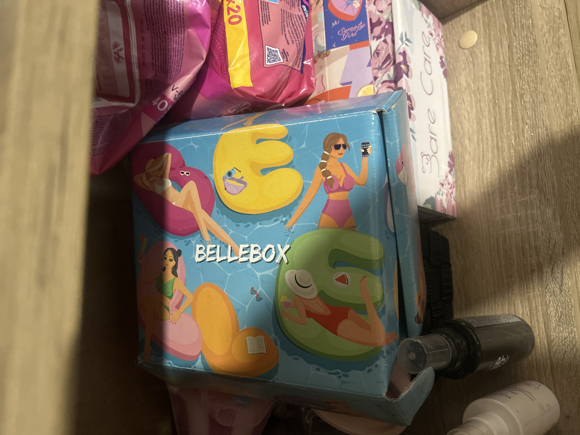The image shows a top-down view of a shelf in what appears to be a storage area. The main object in focus is a blue box with cartoon illustrations of women on inflatable pool toys. The box has the text "BELLEBOX" printed on its side. To the right of the blue box are several pink packages, one of which has the text "40" and "K20" visible. Below the blue box, a box with the text "Bare Care" and floral patterns is visible. In the foreground, there are two small bottles, one white and one black. The shelf is made of light-colored wood. The overall environment is cluttered and appears to be indoors. There is no visible indication of the city of Perushtitsa, Bulgaria.