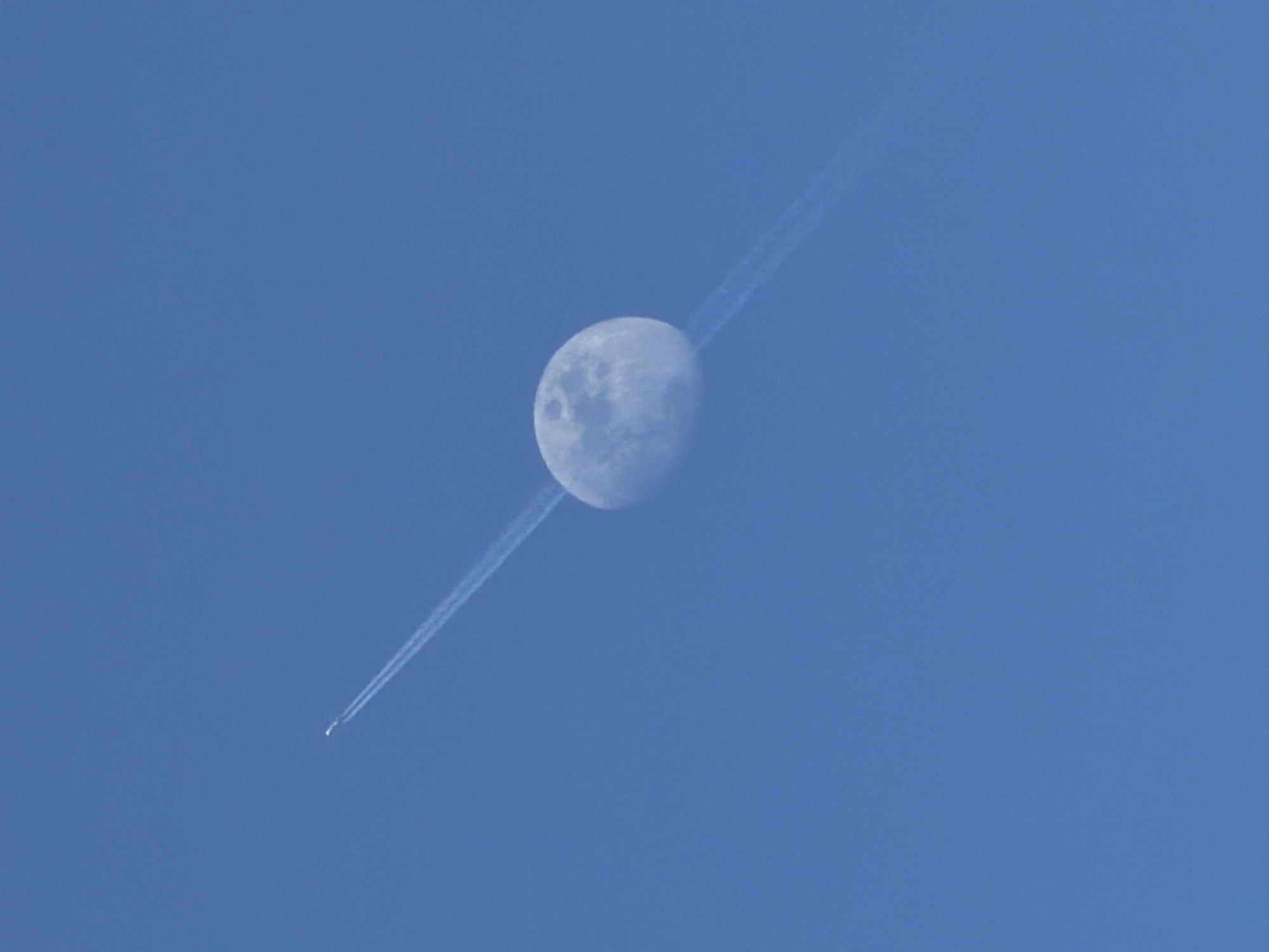 This media file captures a striking celestial scene against a clear blue daytime sky over Priseltsi, Bulgaria. The central focus is a half-illuminated moon, likely in its first or third quarter phase, faintly revealing details of its craters. Diagonally cutting across the entire frame, and passing directly in front of the moon, is a distinct white contrail. This contrail originates from a small airplane, visible as a tiny white speck at the lower-left end of the trail. The airplane and its contrail create a dynamic line that contrasts with the static, ethereal presence of the moon. The sky is a uniform, bright blue, suggesting clear weather conditions. There is no visible text in the image.