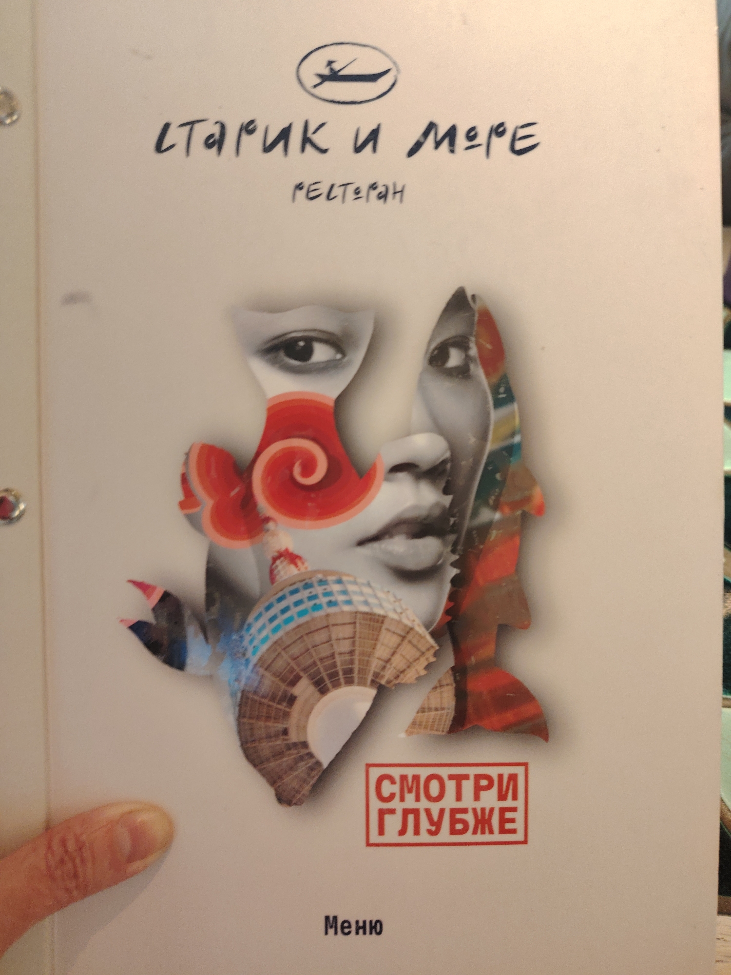 The image shows the front cover of a menu from a Russian restaurant, held by a person whose thumb and partial index finger are visible on the lower left. The menu cover features a complex and artistic collage as its main visual element. This collage intertwines grayscale depictions of human facial features—an eye and lips—with vibrant, abstract shapes in shades of red, blue, and brown. These shapes include a prominent red spiral, a fan-like structure with a mosaic pattern, and a serrated outline reminiscent of a fish.
At the top of the cover, a logo depicts a small boat with a person rowing, enclosed within an oval. Below this, the restaurant's name is written in stylized Cyrillic script: "СТАРИК И МОРЕ" (Starik i More), which translates to "The Old Man and the Sea." Underneath, the word "РЕСТОРАН" (Restoran), meaning "Restaurant," is displayed in smaller text.
A distinctive red square near the bottom center of the cover contains the phrase "СМОТРИ ГЛУБЖЕ" (Smotri Glubzhe), translating to "Look Deeper," which reinforces the intriguing nature of the artwork. At the very bottom, the word "Меню" (Menyu) is printed. The use of Cyrillic script indicates a Russian-speaking context, consistent with the provided location of Moscow, Russia. The menu appears to be bound with metal rings, partially visible on the left edge. The scene is likely indoors, within a restaurant setting.