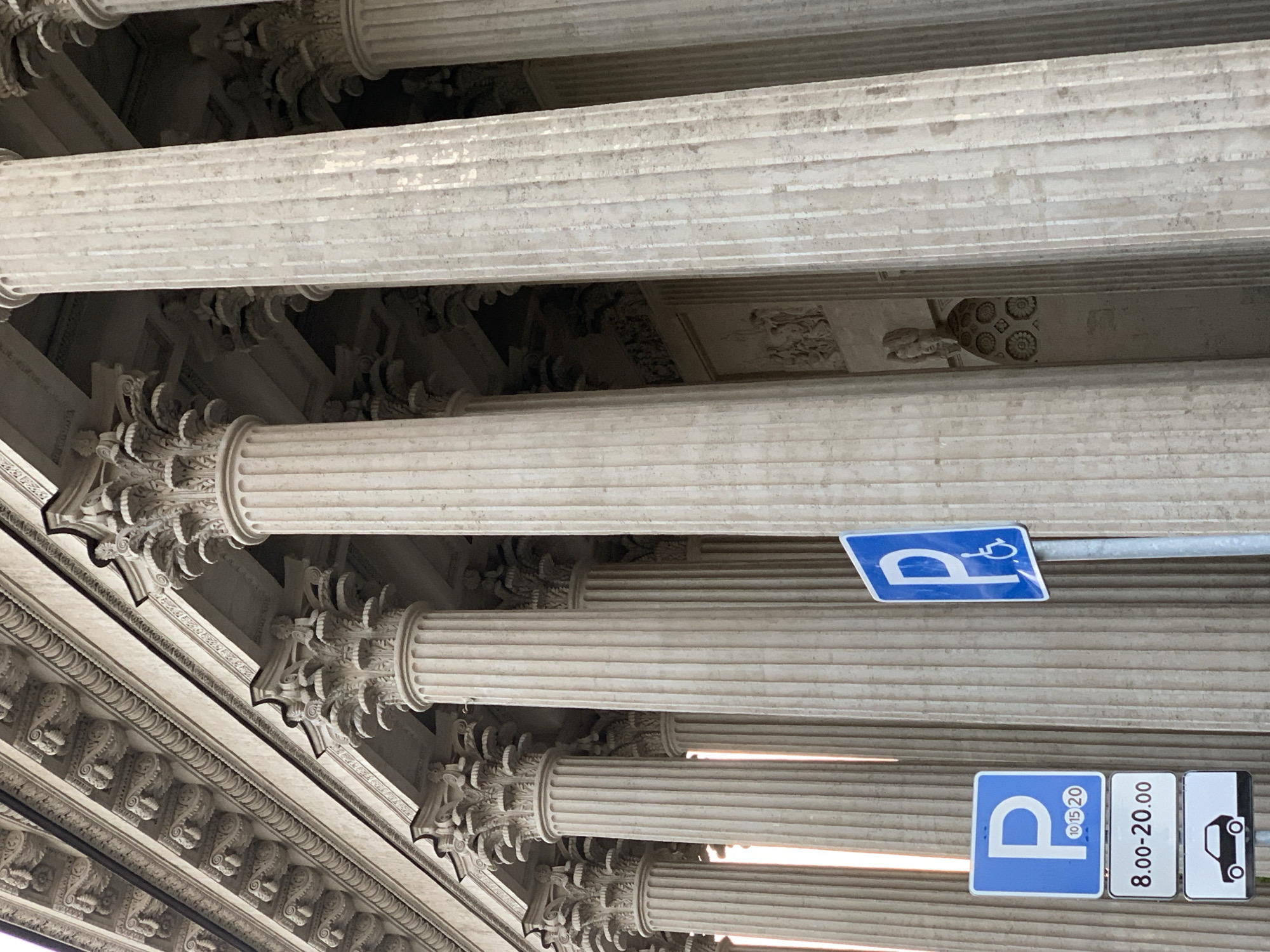 The image shows a low-angle view of a building facade with Corinthian columns, likely located in Saint Petersburg, Russia. The columns are made of light-colored stone or plaster and feature detailed carvings. In the foreground, there are two blue and white parking signs attached to a metal pole. The top sign indicates disabled parking, while the bottom sign shows parking is allowed from 8:00 to 20:00 with 10, 15 and 20 on the P. The background is filled with rows of columns and intricate architectural details of the building's ceiling.