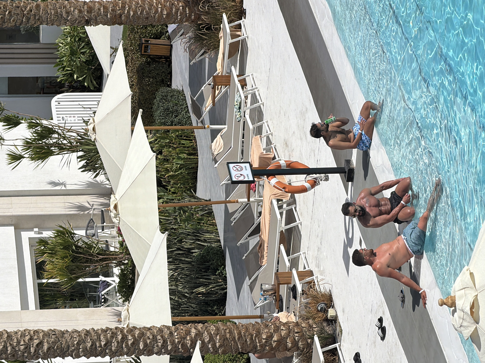 The image depicts a resort swimming pool area in Sharm El Sheikh, Egypt. Three men are present. One man sits on the edge of the pool, with his legs submerged in the water, holding a green can to his lips. Another man, with a muscular physique, is crouched at the edge of the pool. A third man is seated on the pool edge, looking towards the other two men, with his legs in the water. The background features several empty lounge chairs under large white umbrellas, some with towels draped over them. Palm trees and lush green foliage are visible, along with a white building with large windows. The pool water is a vibrant turquoise blue.
