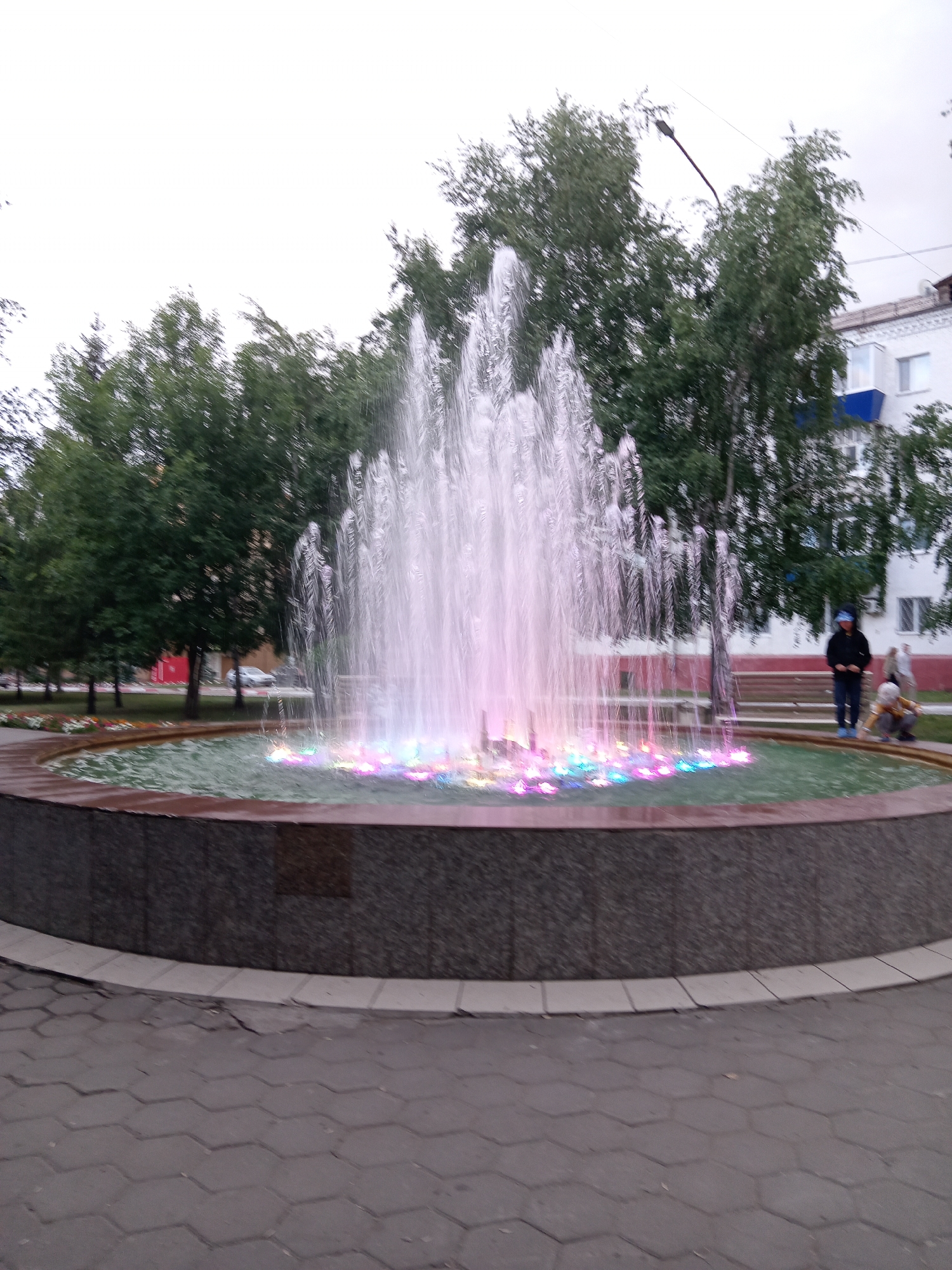 A wide shot captures a lively fountain in a park or public square in Rudny, Kazakhstan. The fountain is the central focus, with water jets shooting upwards and illuminated by vibrant pink, purple, and blue lights at its base. The water's luminescence suggests it's either dusk or evening, or that the fountain is specifically designed for nighttime display.
Around the fountain, a curved, dark-tiled border separates it from the paved walkway. Beyond the fountain, lush green trees form a dense backdrop. To the right, a white building with blue accents and windows is visible, suggesting an urban or residential setting. A lamppost with a curved arm stretches across the upper portion of the frame, hinting at the park's illumination.
Several figures are present. In the middle ground, a person wearing a dark jacket and blue pants stands near a bench, looking towards the fountain. To their right, a child in a light-colored outfit is crouched down, seemingly interacting with something on the ground. Further to the right, another indistinct figure is partially visible. The presence of people and the vibrant fountain suggest a public space that is actively used and enjoyed by the community. The weather appears overcast. No visible text is present in the image.