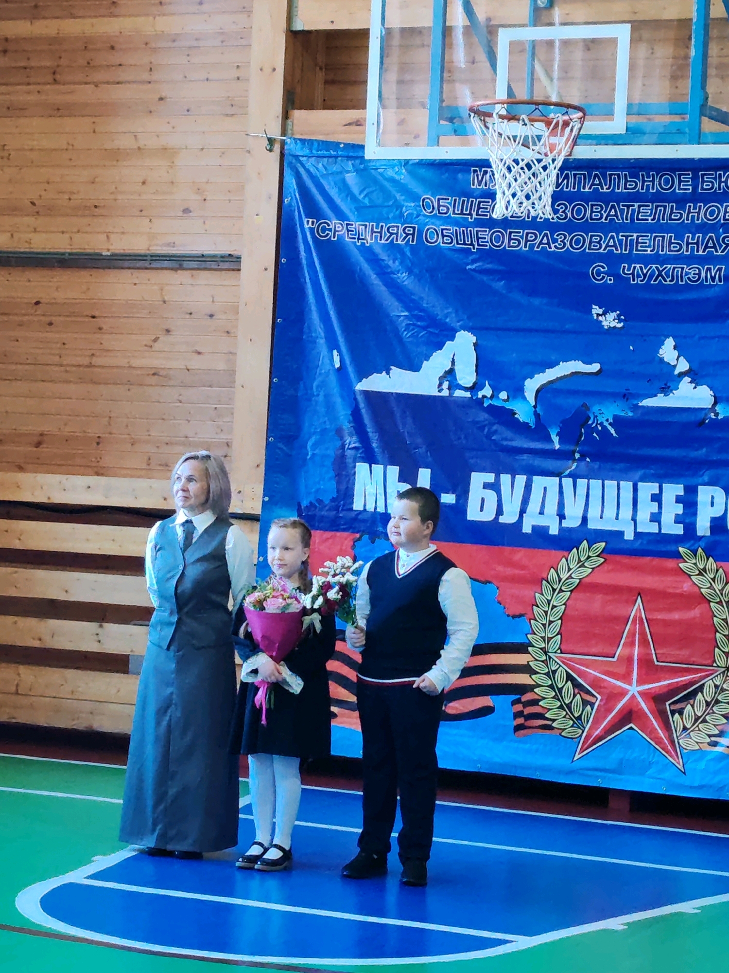 In an indoor gymnasium with wooden paneled walls, three individuals stand on a blue and green court marked with white lines, facing a large blue banner. A basketball hoop with a white net is visible in the upper portion of the frame, positioned behind the banner.
On the left, a woman with short, light brown hair, wearing a dark gray long skirt, a white blouse, a dark gray vest, and a dark tie, stands with her hands clasped behind her back.
In the center, a young girl with light brown hair pulled back, wearing a black dress with white trim on the cuffs and collar, and white tights with black shoes, holds a bouquet of pink and red flowers with a white ribbon.
To the right of the girl, a young boy with short brown hair, wearing a white long-sleeved shirt and a dark V-neck vest, stands with his hands at his sides.
The blue banner behind them features a world map outline in white, and text in white and yellow lettering, some of which reads "МЫ - БУДУЩЕЕ РО" and appears to be in Russian. A large red star surrounded by a green laurel wreath is prominently displayed on the lower right side of the banner, over a background of orange and black stripes, suggesting a connection to Victory Day or patriotic themes. The text on the banner likely indicates the banner is for a municipal educational institution in the city of Чухлэм, Russia.