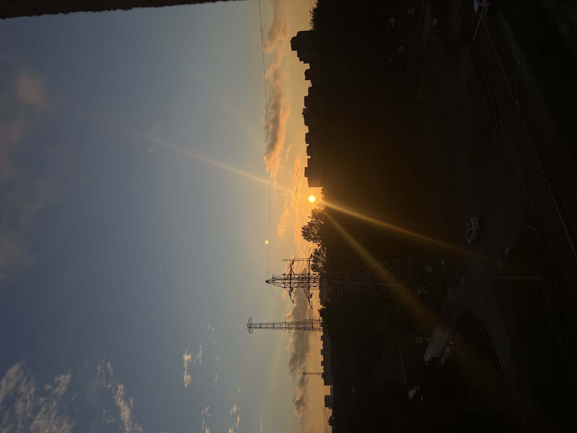The image depicts a sunset in an urban environment, likely Saint Petersburg, Russia, given the provided context. The sun is low on the horizon, casting long rays of light across the scene, creating a warm, orange glow. Wispy clouds are visible in the sky, illuminated by the setting sun.
Dominating the silhouette of the cityscape are several tall, lattice-like structures, indicative of electricity pylons. Behind these, the outlines of apartment buildings are discernible against the bright sky. In the foreground and to the right of the image, a dark asphalt road is visible, with lane markings and a curved intersection. A white vehicle is on the road. Streetlights are also present along the roadside.
The time of day is clearly sunset, characterized by the low sun, the vibrant colors in the sky, and the lengthening shadows. The weather appears clear, with the exception of the scattered clouds. The overall mood is serene and atmospheric, capturing the beauty of an urban sunset. No visible text is present in the image.