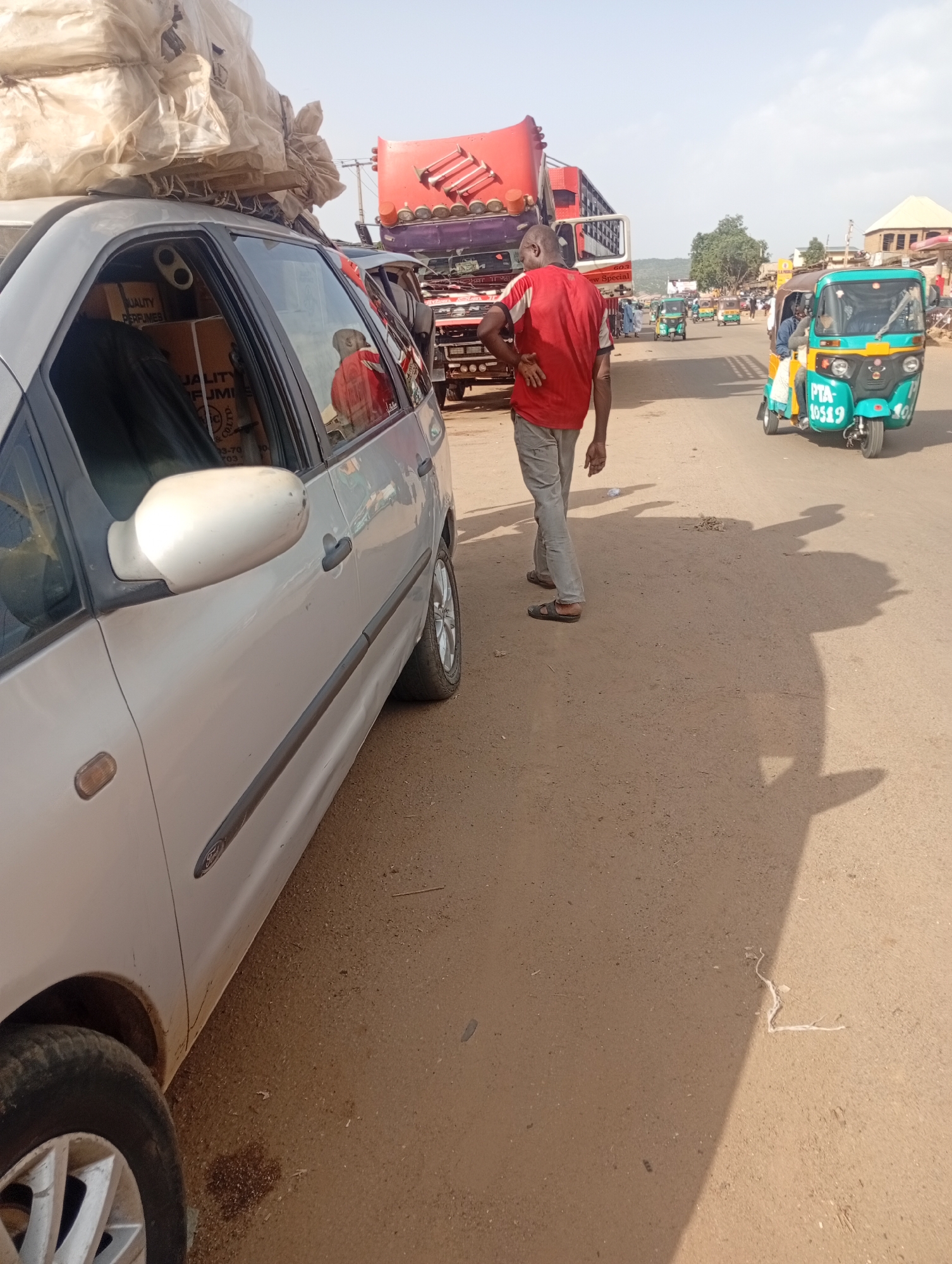 The image depicts a street scene in Rafin Par, Nigeria, likely during the day given the bright sunlight casting long shadows. The foreground is dominated by the side view of a silver car, its roof piled high with a large, light-colored load wrapped in plastic sheeting. Through the open driver's side window, boxes labeled "QUALITY PERFUMES" are visible, suggesting the car is either transporting goods or has recently been loaded.
In the middle ground, a man in a red t-shirt and grey trousers stands facing away from the camera, his hands on his hips, appearing to observe the traffic. Behind him, a large red truck with a white trailer is stopped or moving slowly. Further down the road, several colorful auto-rickshaws, commonly known as "Keke Napep," are visible, some in motion and others parked. One turquoise and yellow auto-rickshaw in the right middle ground displays the license plate "PTA 10519". The overall setting appears to be a dusty, possibly unpaved or partially paved road with some buildings and vegetation in the background, indicative of a bustling market or roadside area. The atmosphere seems active and commercial, typical of a busy transportation hub.
