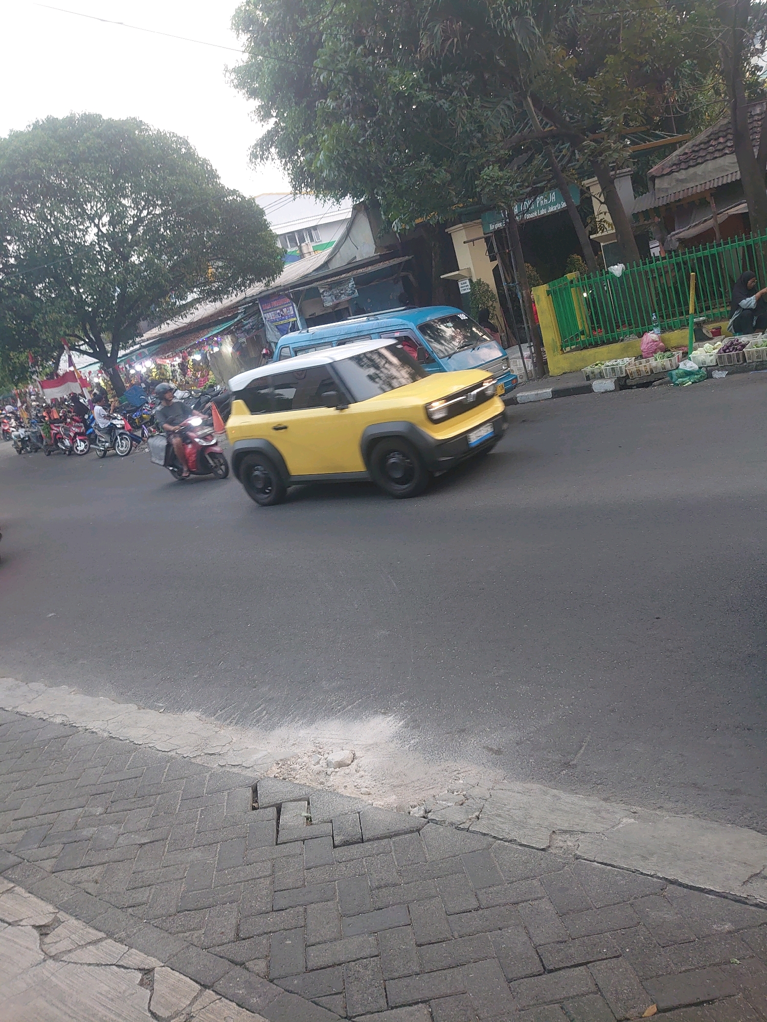 The image depicts a street scene in South Jakarta, Indonesia. In the foreground, a yellow compact car is captured in motion, moving from left to right across the asphalt road. To the left of the car, several motorcycles are parked or moving, indicating a bustling area. Further to the left, market stalls are visible, adorned with colorful decorations, suggesting a commercial or market district.
Behind the yellow car, a blue van is also present on the road, partially obscured by the car. The background reveals trees, some with lush green foliage, and buildings that appear to be shops or residences. A green metal fence runs along the right side of the image, separating the street from a sidewalk area where several baskets filled with produce, possibly vegetables, are displayed. A person, likely a vendor, is partially visible near the fence, interacting with the goods.
The pavement on the left side of the road is made of interlocking gray bricks, showing some signs of wear and tear with cracks and unevenness. The overall environment suggests a typical urban street in Indonesia, characterized by vehicle traffic, market activities, and pedestrian walkways.