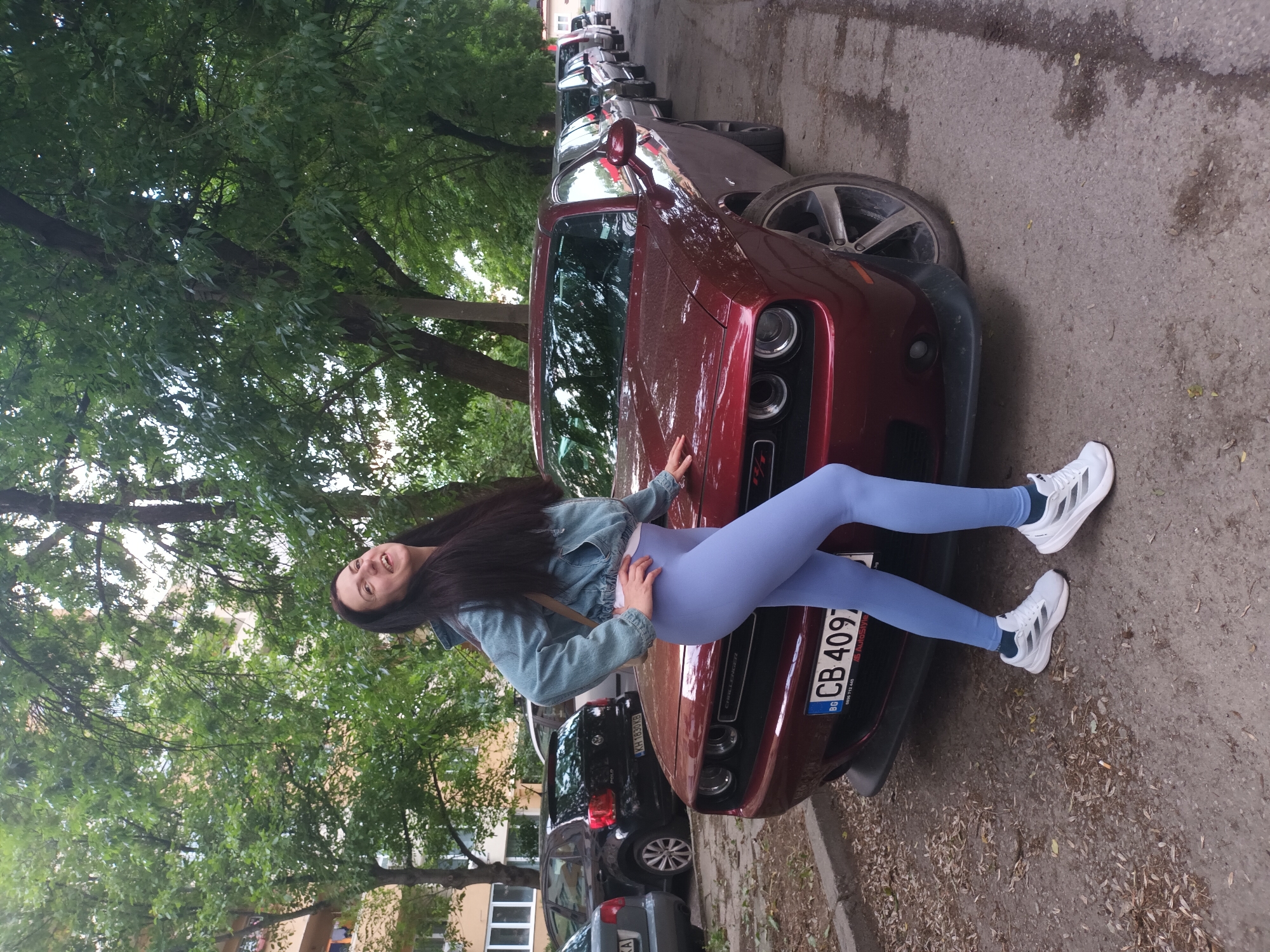 A young woman in blue leggings and a denim jacket reclines on the hood of a maroon Dodge Challenger parked on a city street. The vehicle has license plate "CB 4097." Several cars are parked behind the Challenger, and trees line the street. The location is identified as an unknown city, Unknown.