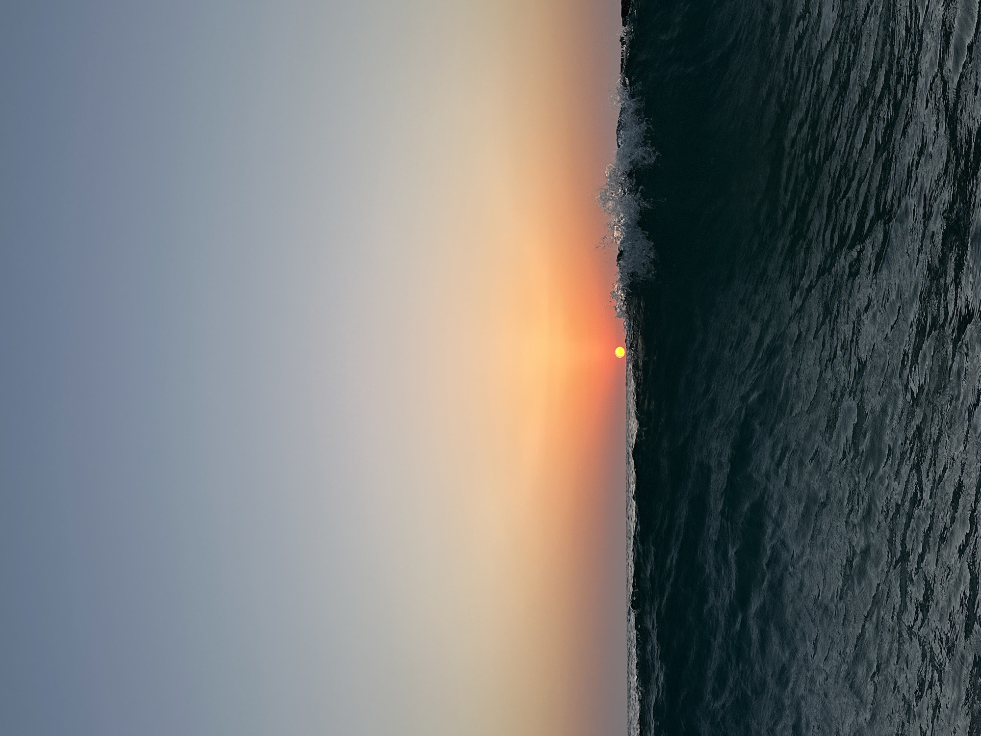 The image depicts a sunset over the ocean. A large wave is breaking in the foreground, with foam and spray visible at its crest. The sun, a bright orange orb, is partially obscured by the wave as it sets on the horizon. The sky above transitions from a pale blue at the top to shades of orange and pink closer to the horizon, indicating the time of sunset. The water is a deep, dark blue-green, with visible ripples and texture. The scene captures a dynamic moment of the ocean meeting the setting sun. Given the context of Tel-Aviv, Israel, this scene would likely be observed from one of the city's beaches, such as Gordon Beach or Frishman Beach, facing west towards the Mediterranean Sea. There are no people or man-made structures clearly visible in the frame.