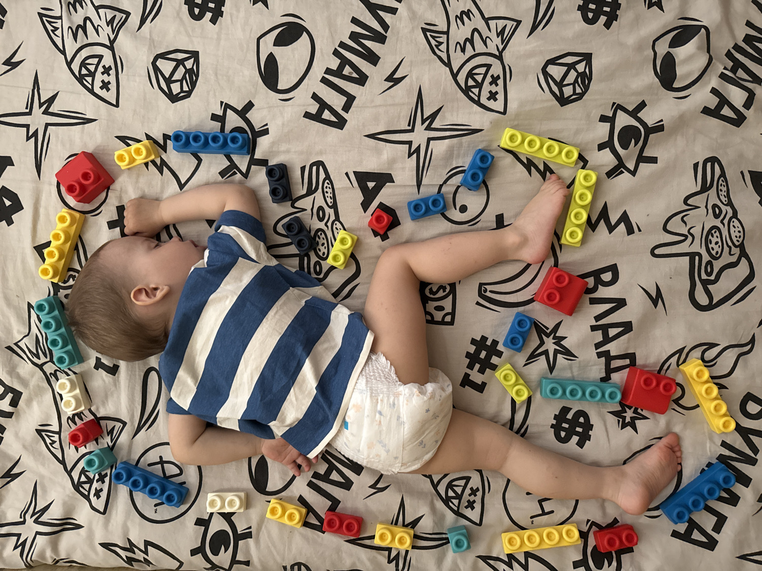 A high-angle overhead shot captures a baby or toddler peacefully sleeping on a bed or soft surface, surrounded by colorful building blocks. The child lies on their right side, facing to the left, with their right arm bent and hand resting near their face. They are dressed in a blue and white striped short-sleeved t-shirt and a white disposable diaper.
The surface beneath the child is a white sheet or blanket adorned with various black, stylized graphics. These illustrations depict items such as a fish, an alien head, a geometric shape, a dollar sign, a banana, a starburst, a slice of pizza, a lightning bolt, a hashtag, and an eye symbol. Visible text on the sheet includes the Cyrillic word "УМАТА" (UMATA) and the repeated letters "BAAA."
Numerous building blocks, similar to Lego Duplo but appearing to be soft-material, are scattered across the sheet and around the child. These blocks come in red, yellow, blue, and teal colors, featuring various configurations of studs. The overall scene suggests an indoor setting with bright, even lighting, and the child appears calm and asleep amidst their toys.