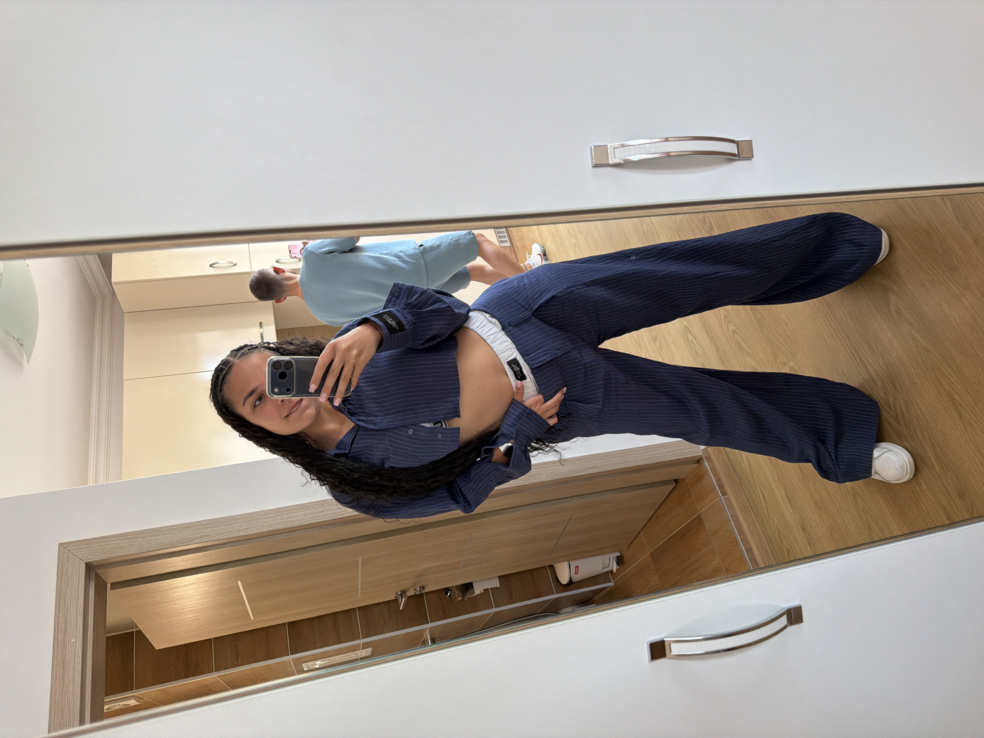 A young woman with braided dark hair takes a mirror selfie in a room with light-colored walls and wooden floors. She is wearing a navy blue pinstripe cropped button-down shirt and matching wide-leg trousers with a white waistband. A man in a light blue t-shirt and shorts is visible in the reflection behind her, looking away from the camera. In the reflection of the mirror to her left, a bathroom with tiled walls and a white trash can can be seen through an open doorway. The scene appears to be an interior, possibly a home or hotel room, located in Nesebar, Bulgaria.