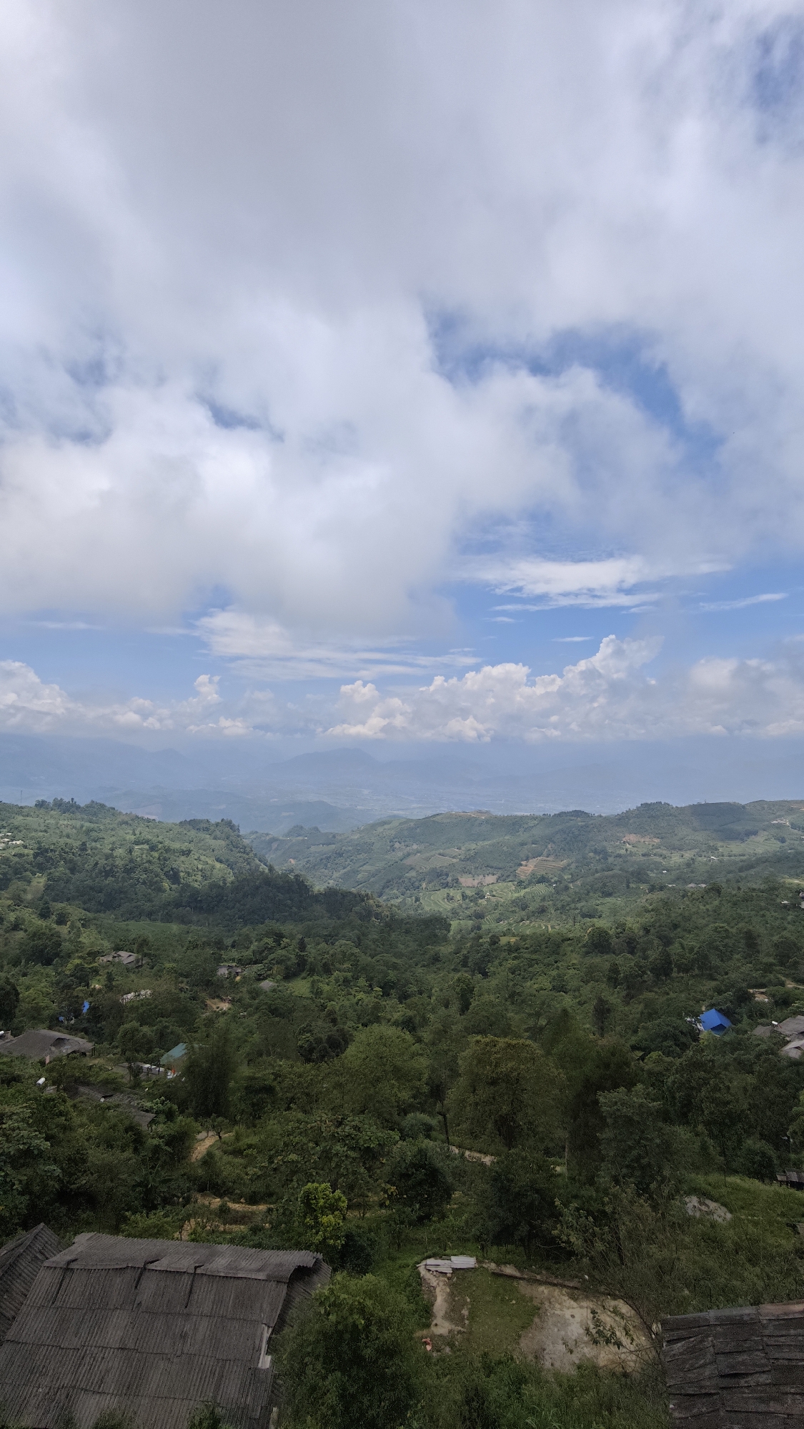 The scene is a view from a high point in the Suối Giàng area of Vietnam. The landscape is dominated by lush, green hills and valleys. Some areas are terraced, likely for agricultural purposes. Scattered throughout the landscape are small structures, probably dwellings, some with dark-colored roofs and one with a blue roof. The sky is partly cloudy with patches of blue, obscuring distant mountains or features on the horizon. The perspective is from above, looking down onto the landscape. Some roofing material from nearby structures is visible in the immediate foreground.