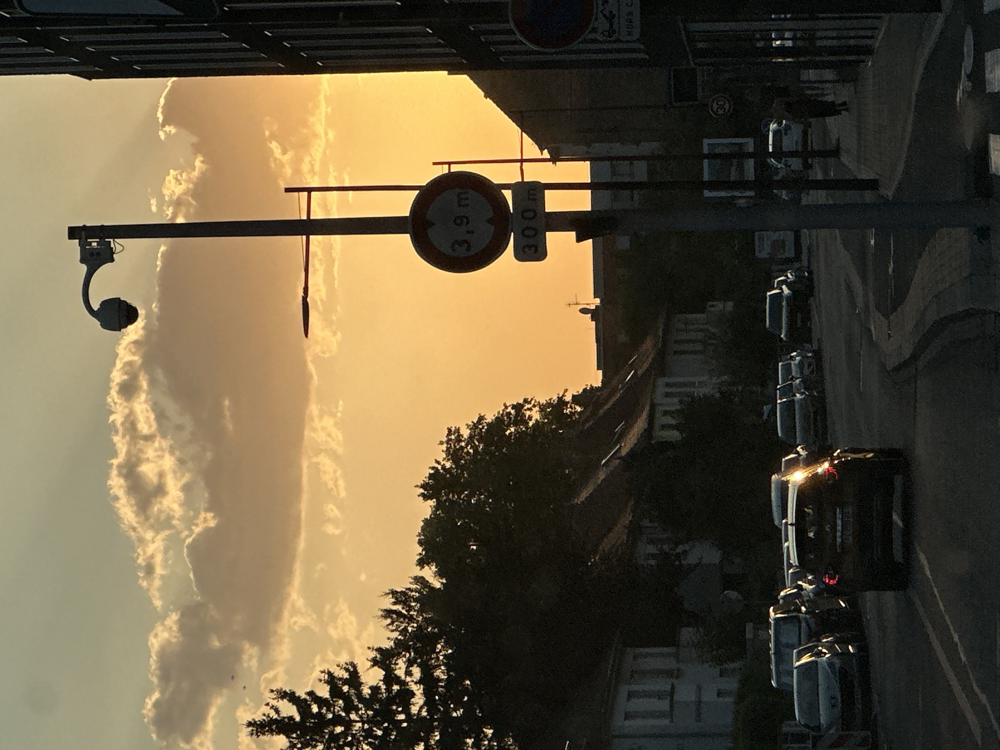 The photograph depicts a street scene at dusk in Strasbourg, France. The sky is filled with golden hues of sunset and dramatic, illuminated clouds, with sun rays piercing through them. In the foreground, several cars are parked and driving on a road. A prominent traffic sign indicates a height restriction of 3.9 meters and a distance of 300 meters. Another circular traffic sign with the number "30" is visible further down the street. On the right side of the image, a dark building with vertical lines and windows dominates the frame. A lamppost with a surveillance camera is also present. On the left side, residential buildings with pitched roofs and trees line the street. A lone figure is walking on the sidewalk in the distance on the right.