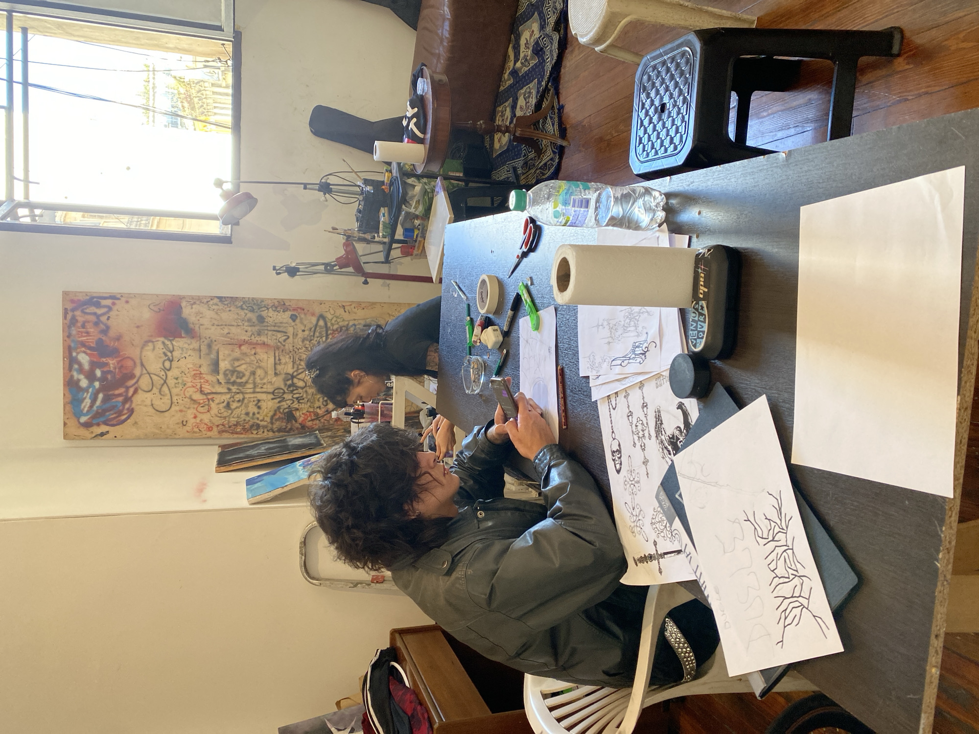 This image captures an indoor scene within a creative studio or workspace in Buenos Aires, Argentina, during the daytime. Bright sunlight floods the room through a large window at the top of the frame.
Two individuals are seated at a dark-colored table, which serves as the central focus. In the foreground, a person with dark, curly hair, wearing a dark jacket, is engrossed in their smartphone. Behind them, another person with long, dark hair is leaning over the table, seemingly engaged in an artistic activity.
The tabletop is covered with an assortment of art supplies, including scissors, a tape dispenser, markers, a pencil, an eraser, and two rolls of white paper. A clear plastic bottle of "Villa del Sur" water and a glass are also present. Numerous sketches and drawings are scattered across the table, showcasing intricate designs like skulls, chandeliers, sword-like motifs, and stylized branch patterns. One drawing clearly shows the text "333" and "DELSE" alongside tree-like imagery. A black device, partially visible with the label "VENUS JOUR," sits near the sketches.
In the background, elements of the room include a brown leather sofa, a patterned rug on a wooden floor, and a black guitar case leaning against a wall. An abstract artwork with graffiti-like elements in red, blue, and black adorns the wall, suggesting an artistic atmosphere. The overall scene depicts a calm and focused creative session.