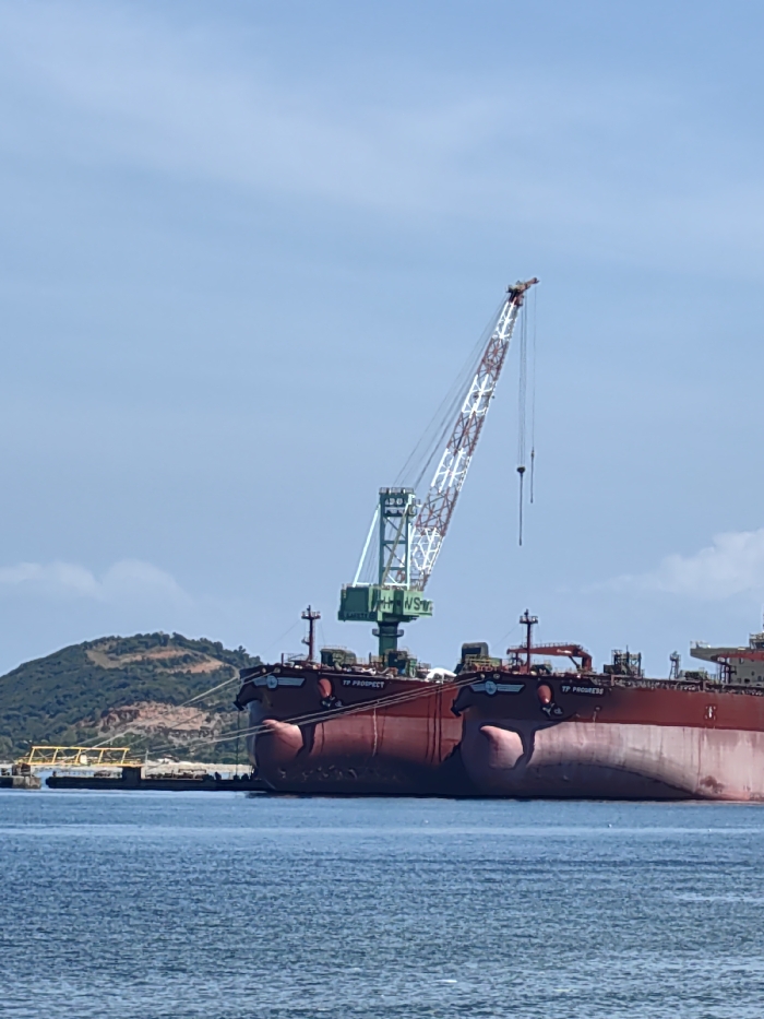 Two red oil tanker ships, named "TP Prospect" and "TP Progress", are docked in a blue body of water. A tall crane with a white and red lattice boom and a green base rises from the ships. The sky is blue and clear. A green hill with brown patches is visible in the background near the dock.