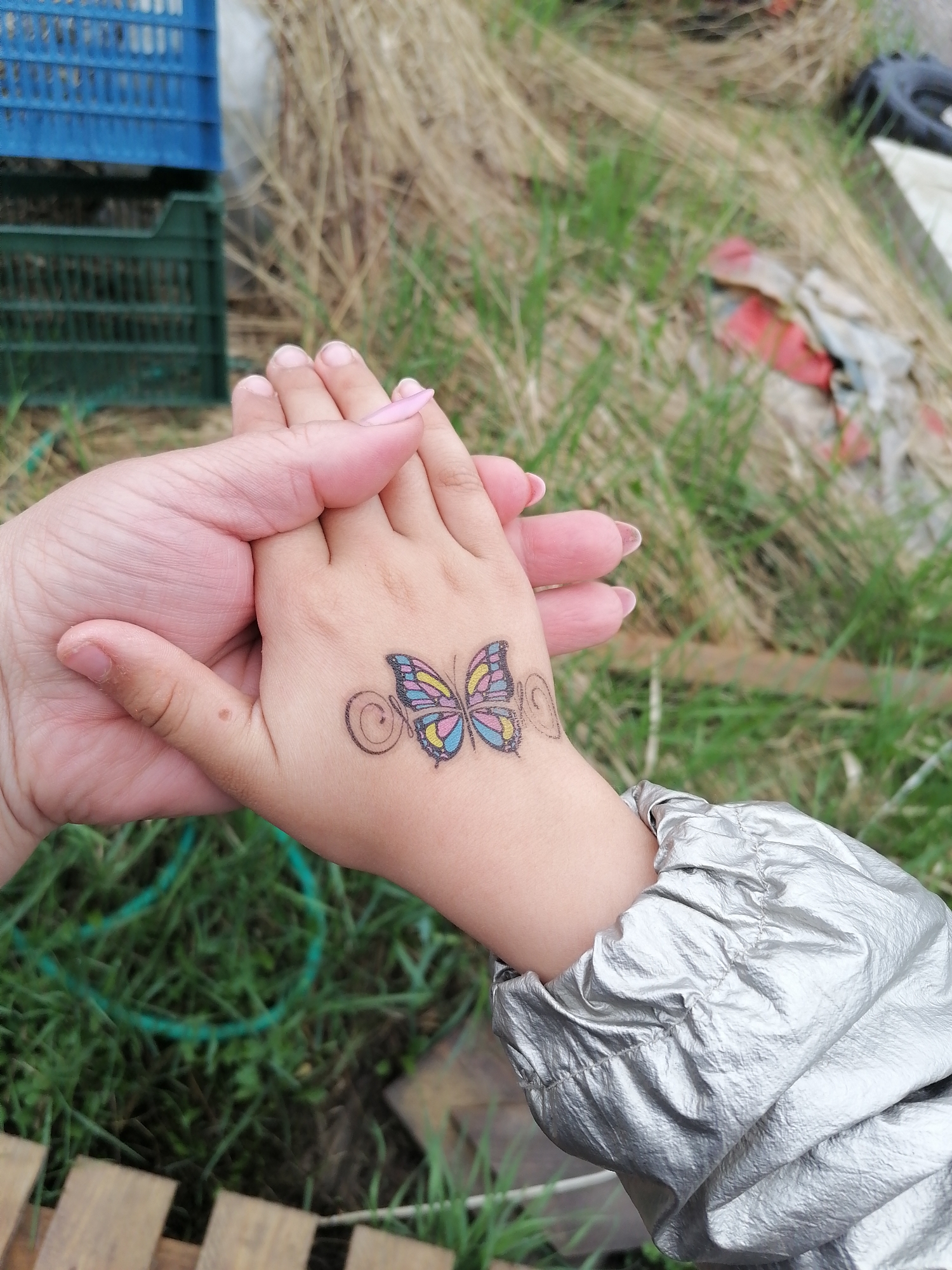 This close-up shot features two hands clasped together, conveying a sense of connection or affection. An adult's hand, with light pink nail polish, gently holds the smaller hand of a child. The child's hand displays a colorful temporary tattoo of a butterfly with pink, blue, and yellow wings outlined in black, flanked by decorative black swirls. The child's arm is partially visible, clad in a crinkled, metallic silver or grey jacket sleeve.
The setting appears to be outdoors, with a natural, somewhat unkempt background. Visible elements include patches of dry, straw-like grass mixed with areas of greener grass. In the upper left background, stacked blue and green plastic utility crates are discernible. Further into the background, indistinct items such as a red and white piece of fabric and a dark, rounded object (possibly part of a tire) are partially visible amidst the grass. A wooden plank structure is subtly present in the bottom foreground. The lighting suggests it is daytime, with no specific weather conditions evident. There is no visible text in the image.
