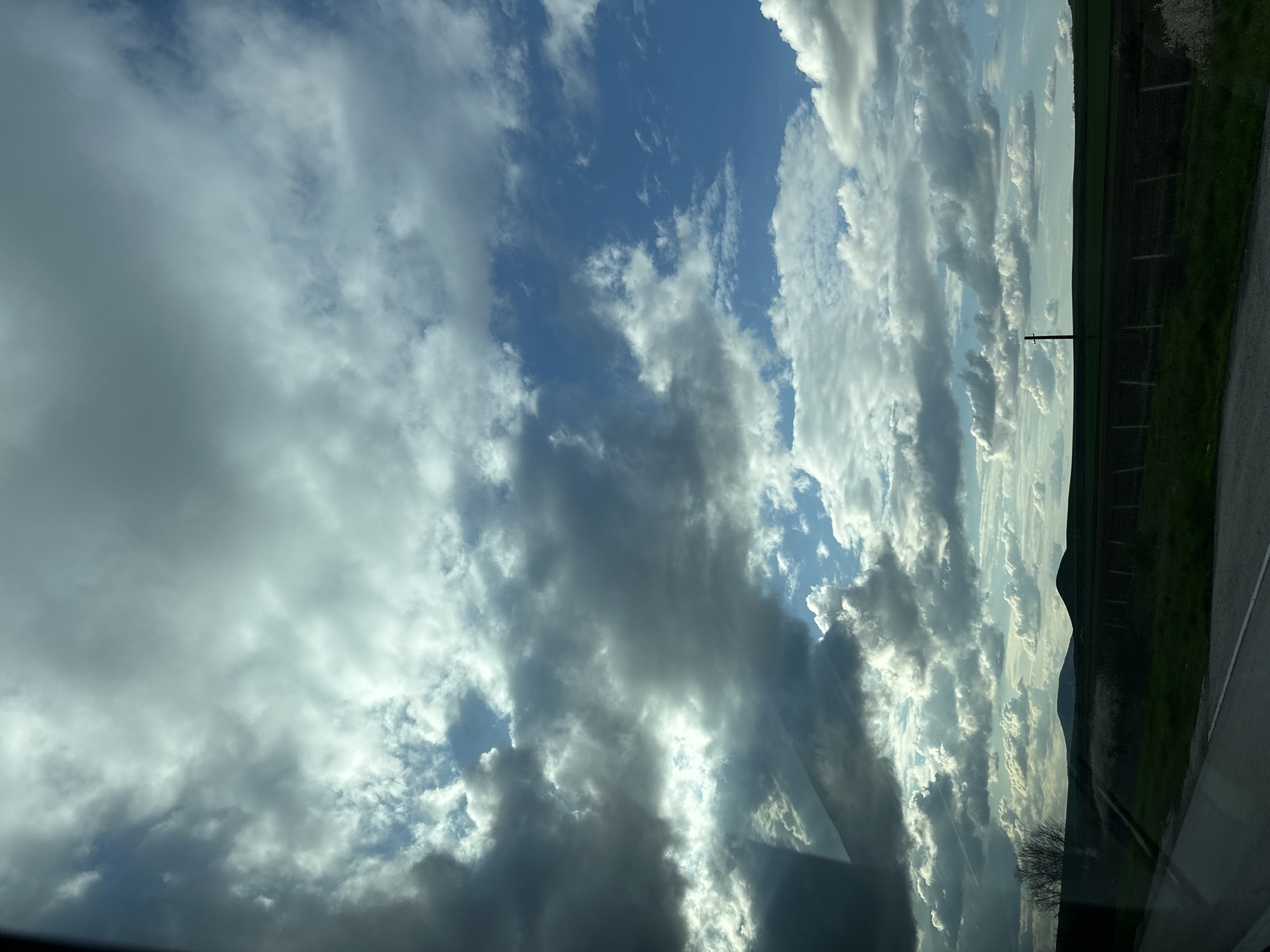 The image presents a view of a sky filled with varying cloud formations, ranging from dense, grey clouds to thinner, lighter ones, against a backdrop of blue sky. The clouds appear to be illuminated by sunlight. Along the horizon, there is a strip of green vegetation and a fence, suggesting a rural or suburban setting. A utility pole is visible against the vegetation. The image is taken from what seems to be an interior perspective, as indicated by a portion of a car or vehicle interior visible at the bottom. While the image captures the sky and landscape, specific information that ties it to the city of Zlatina, Bulgaria, is missing, and the image does not include any people, actions, or interactions.